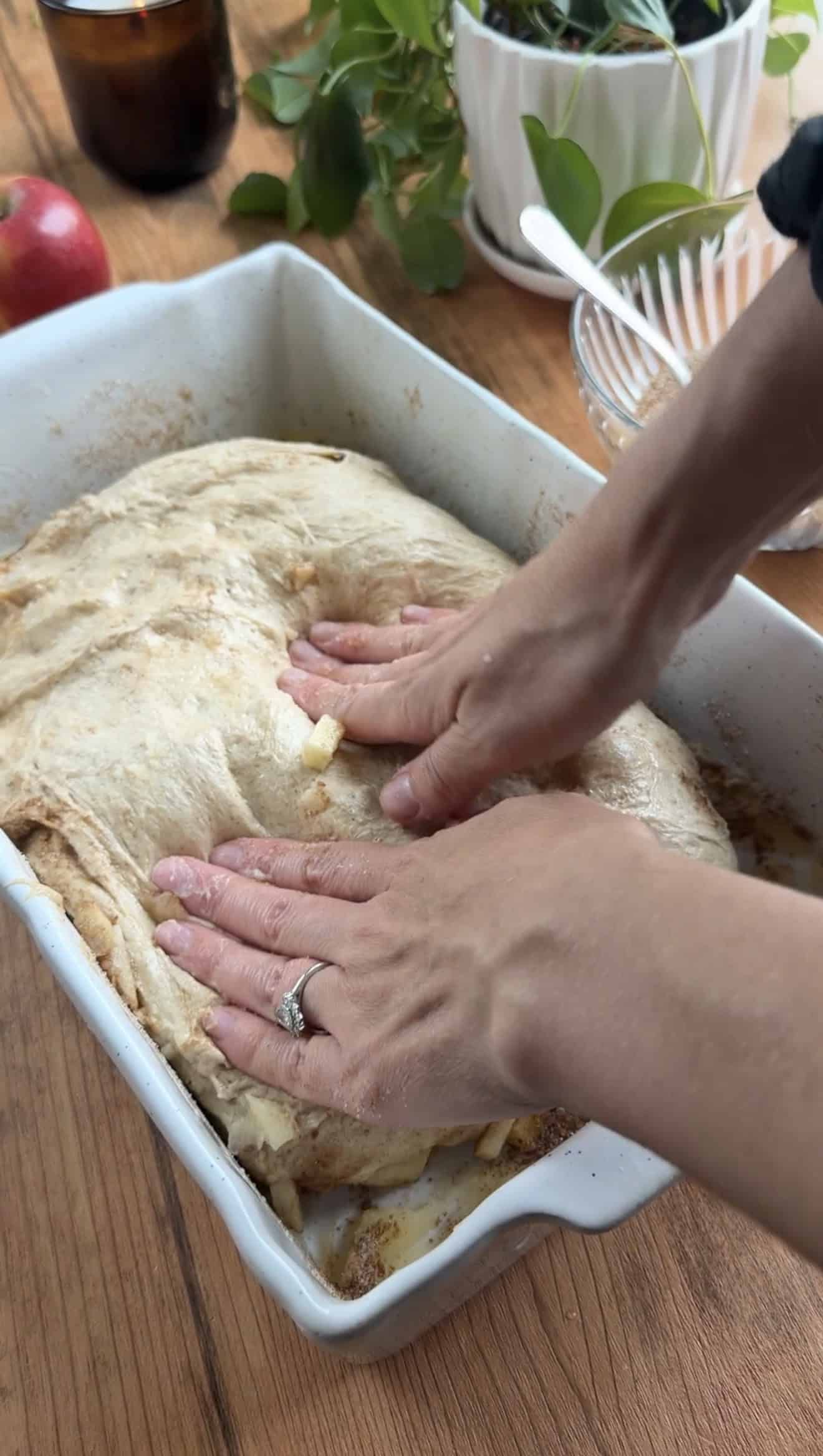 Two hands pressing focaccia dough into the bottom of a white baking dish on a table.
