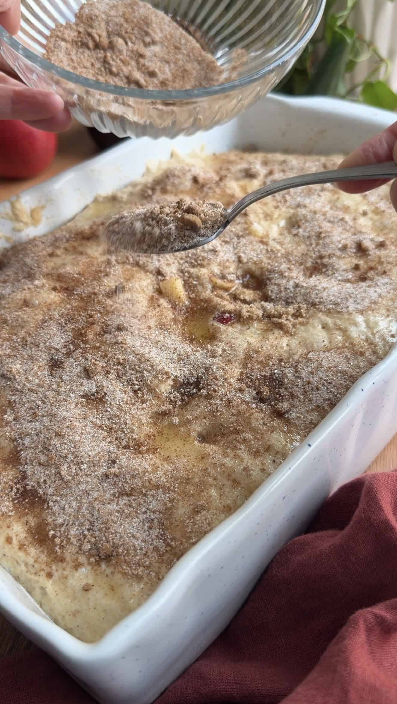 Cinnamon sugar being sprinkled from a metal spoon onto puffy focaccia dough in a white baking dish.