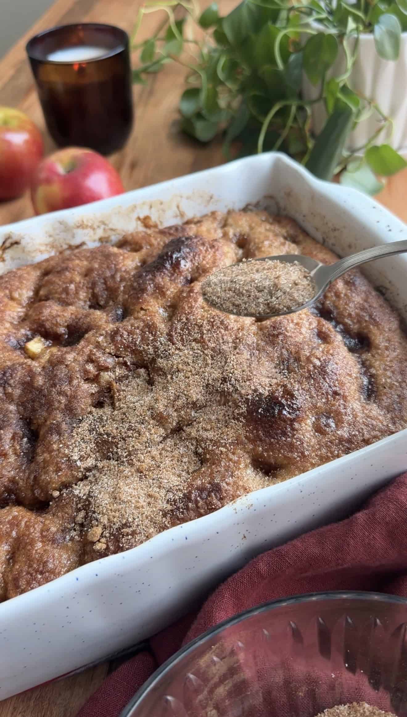 A hand sprinkling cinnamon sugar from a spoon onto baked apple focaccia in a baking dish.