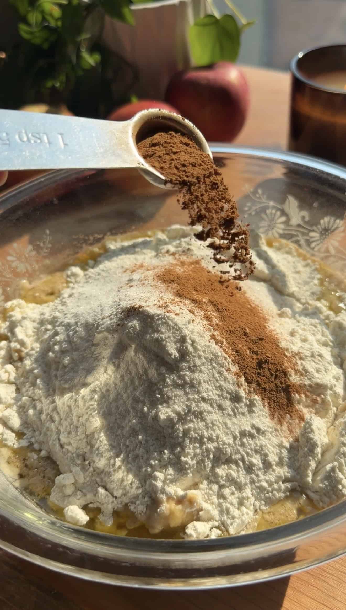 A hand sprinkling cinnamon from a measuring spoon into a glass bowl filled with flour.