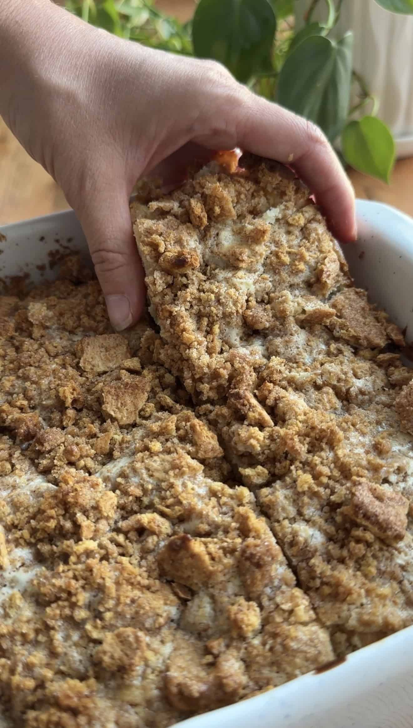 A hand picking up a large piece of cheesecake focaccia from a baking dish filled with more golden bread.