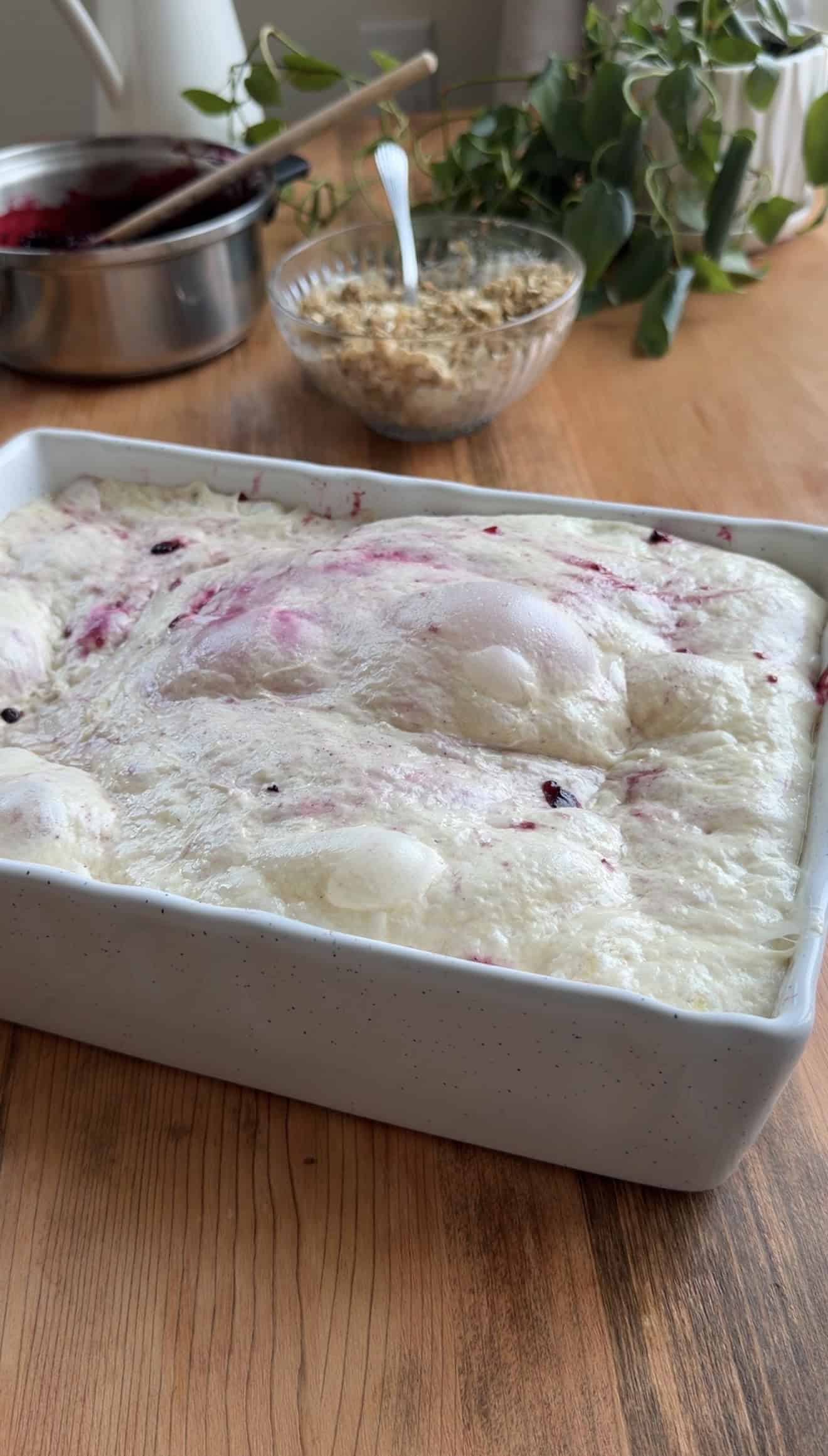Bubbly, proofed focaccia dough in a white casserole dish on a wood table next to a saucepan, plant, and bowl filled with sugar and oats.