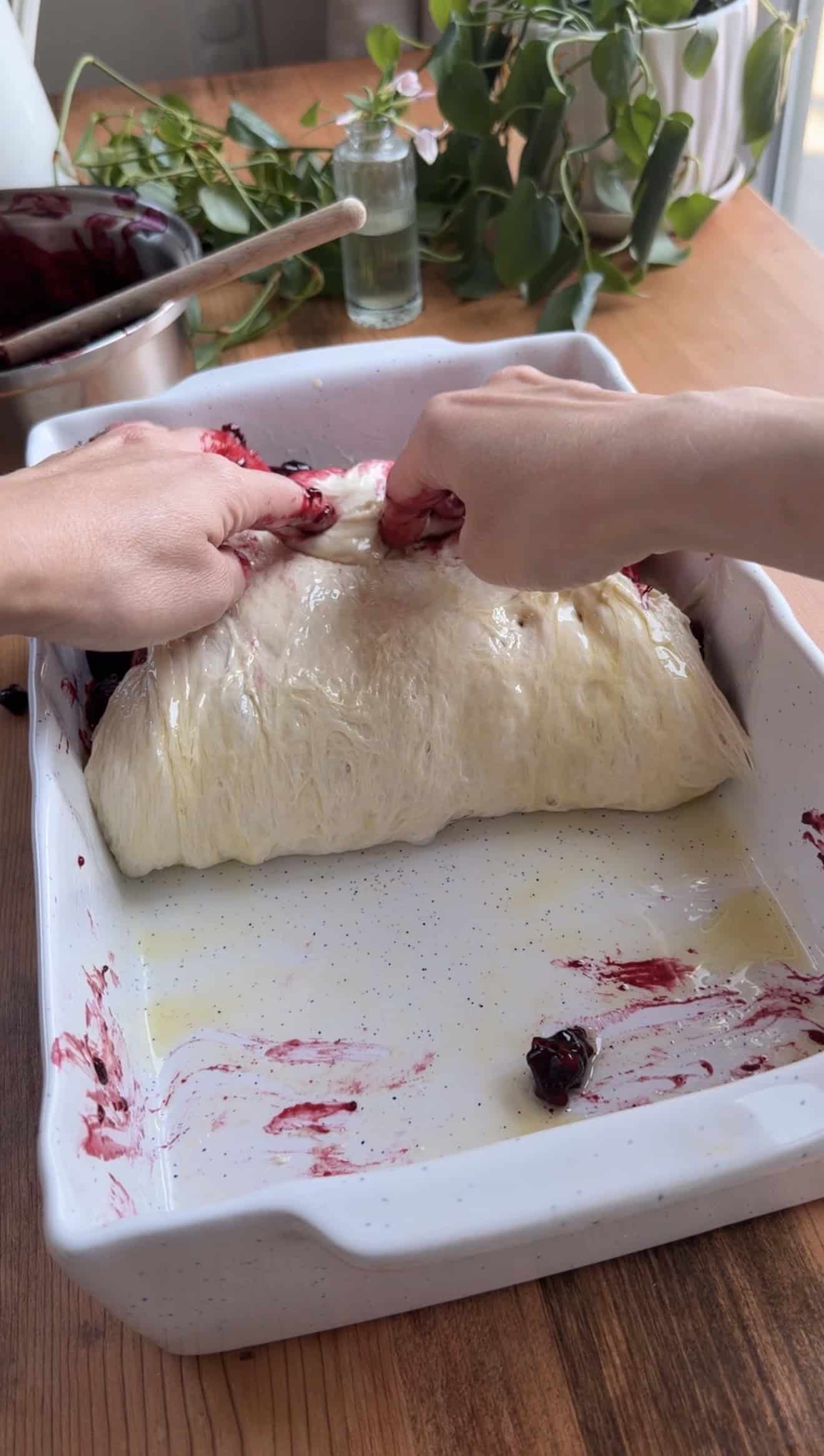 Two hands folding focaccia dough in half, filled with cooked berries, in a baking dish.