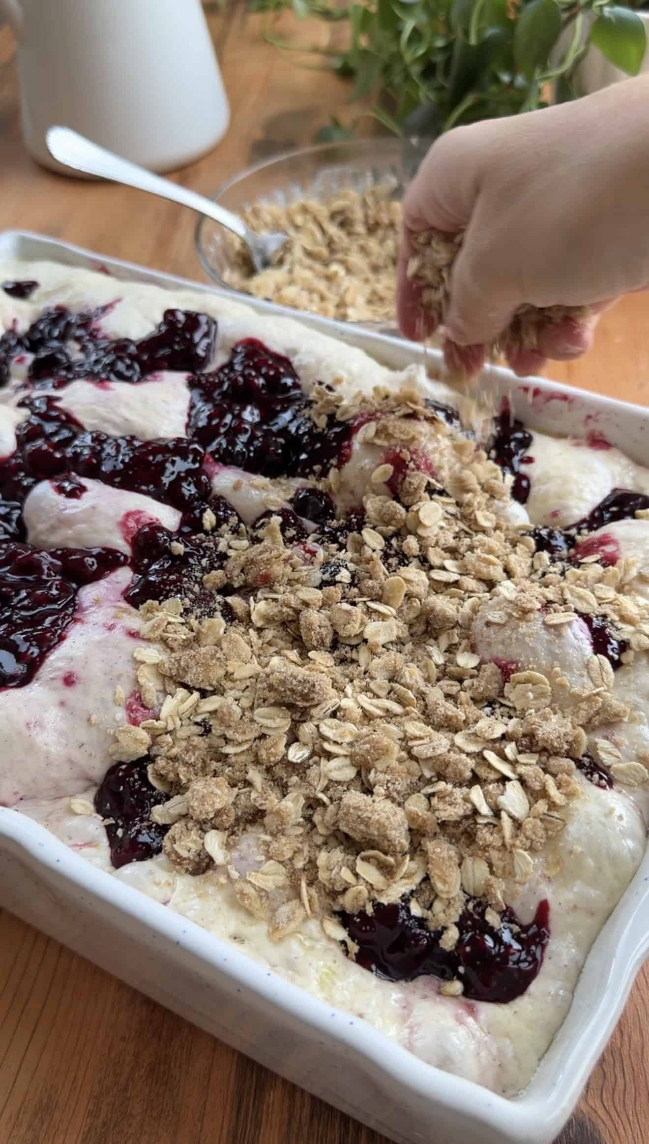 A hand sprinkling crumbly oatmeal streusel onto bubbly focaccia dough that's already topped with cooked berry pie filling in a white baking dish.