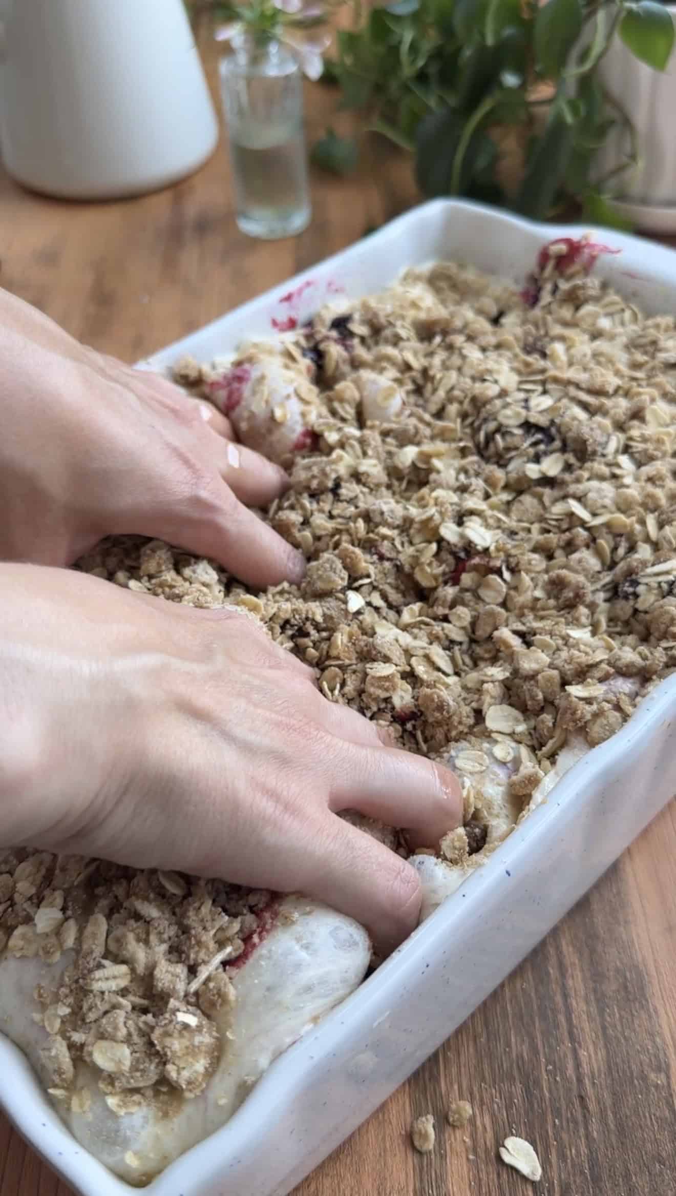 Two hands dimpling focaccia dough in a white baking dish that's topped with cooked blueberries and a mixture of oats, sugar, and cinnamon.