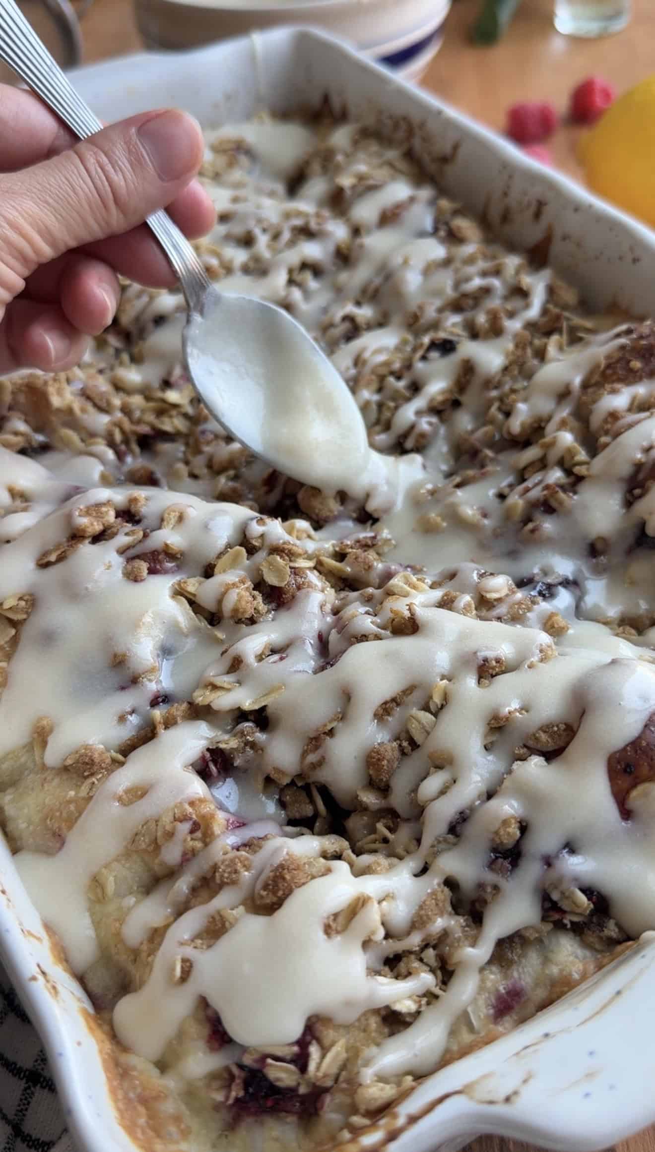 A hand drizzling white glaze from a metal spoon onto baked berry focaccia that's topped with oatmeal streusel.