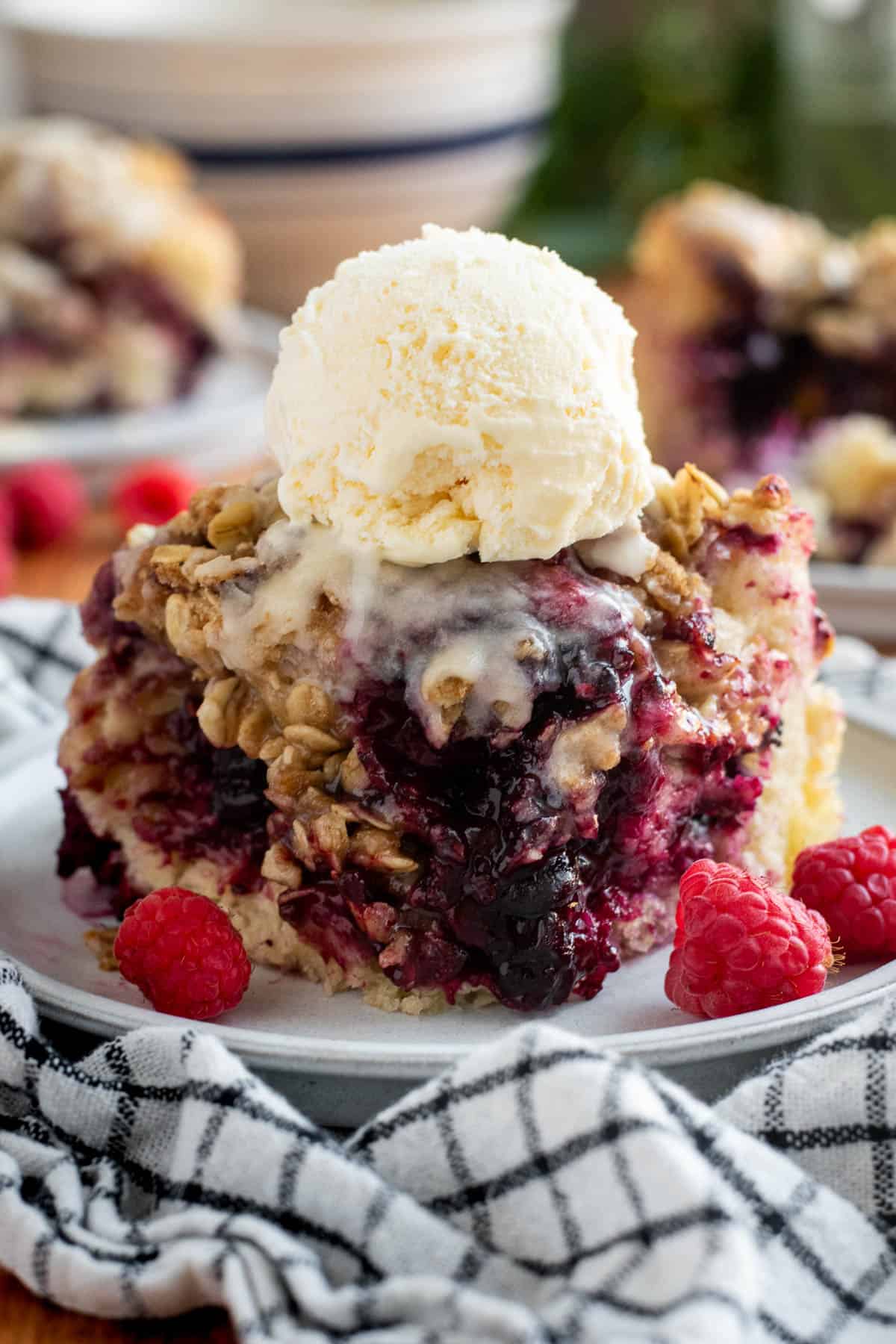 A large piece of berry focaccia topped with glaze, a scoop of vanilla ice cream, and oat streusel on a plate next to three raspberries.