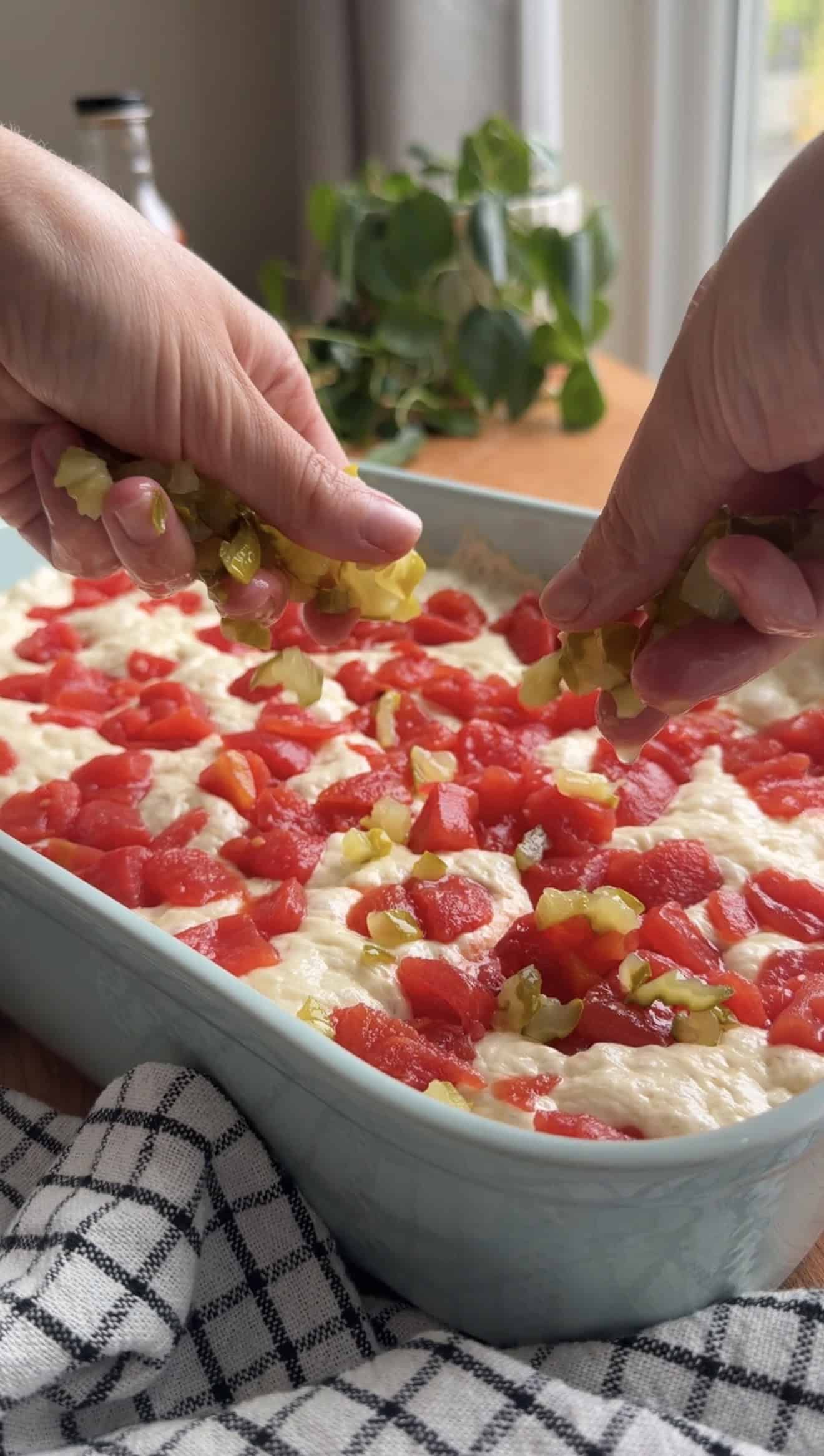 Two hands sprinkling chopped pickles on focaccia dough with diced tomatoes spread over the top of the dough in a baking dish.