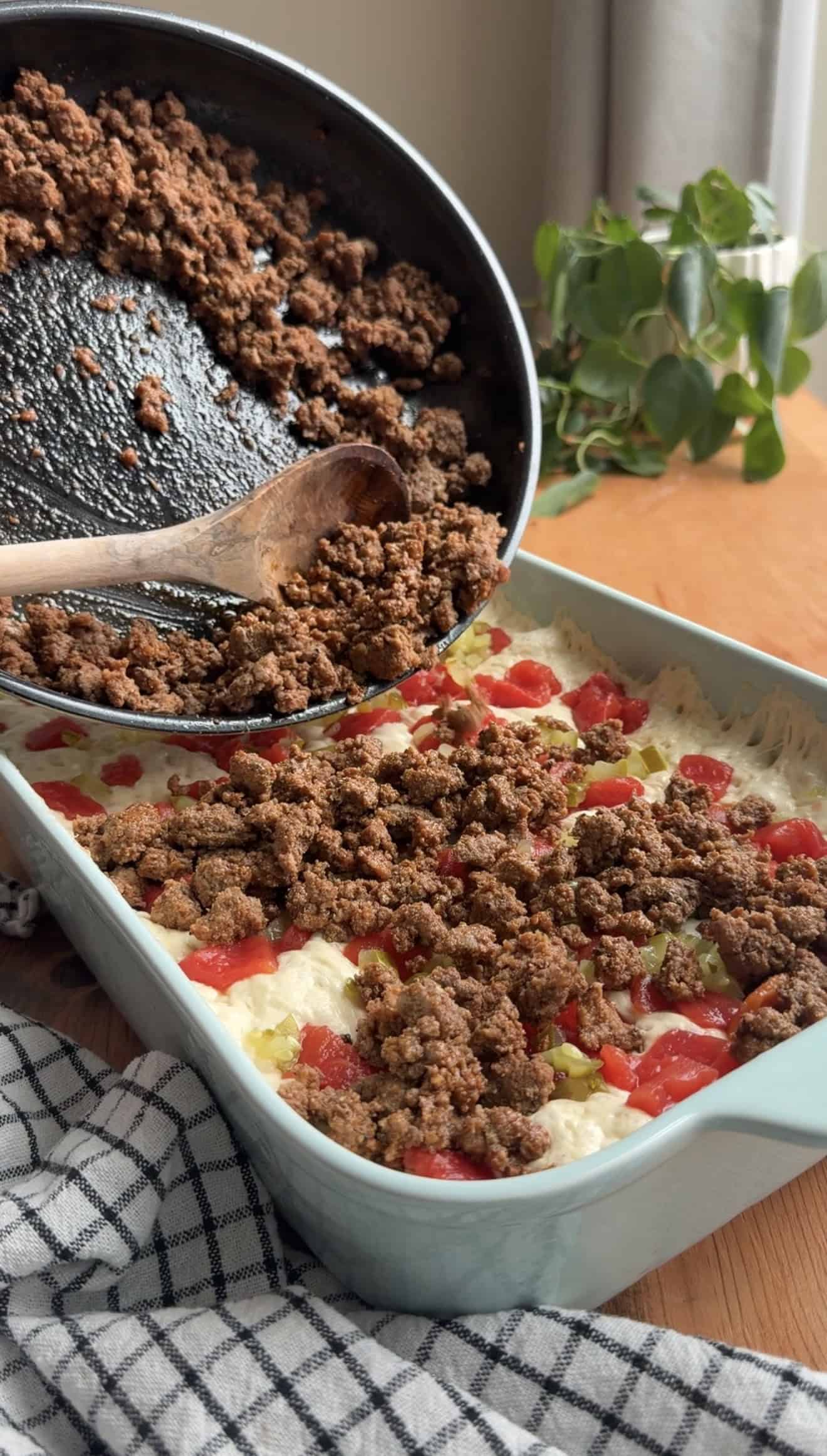 Cooked ground beef being poured from a large skillet onto focaccia dough in a blue casserole dish.