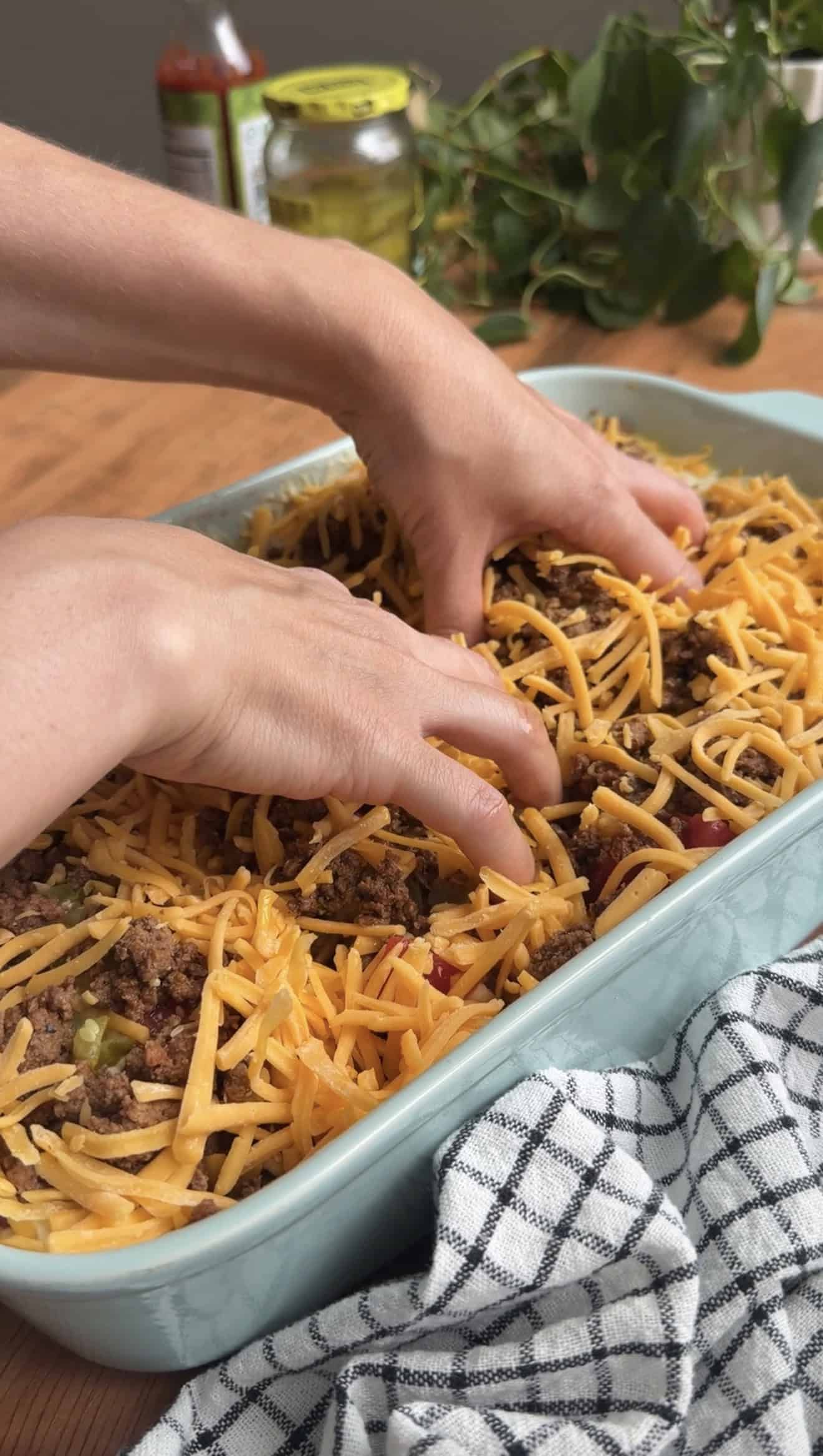 Two hands dimpling focaccia dough in a baking dish topped with browned ground beef and shredded cheddar cheese.