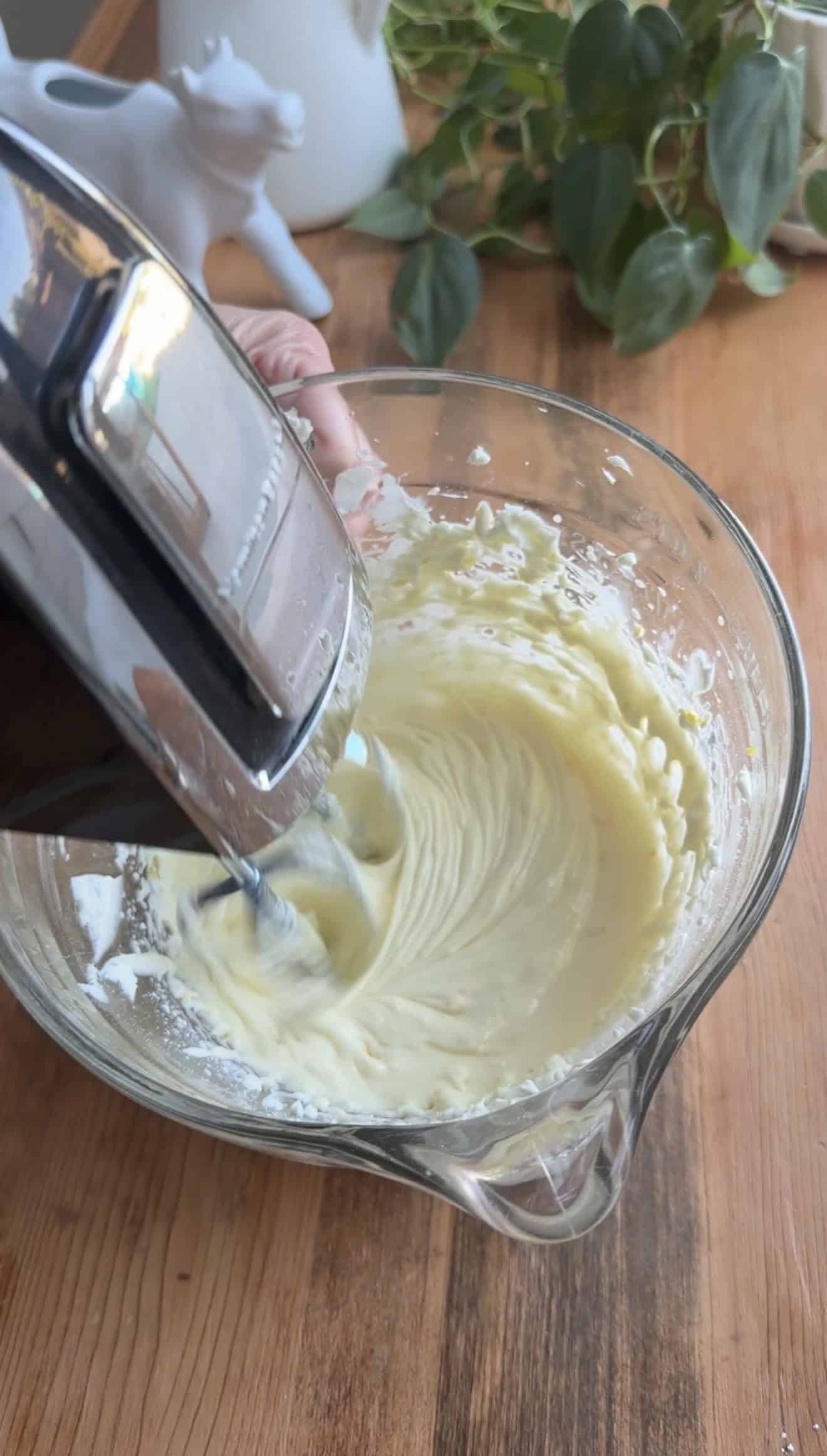 A hand holding an electric hand mixer in a glass bowl of whipped cream cheese, sugar, and egg.