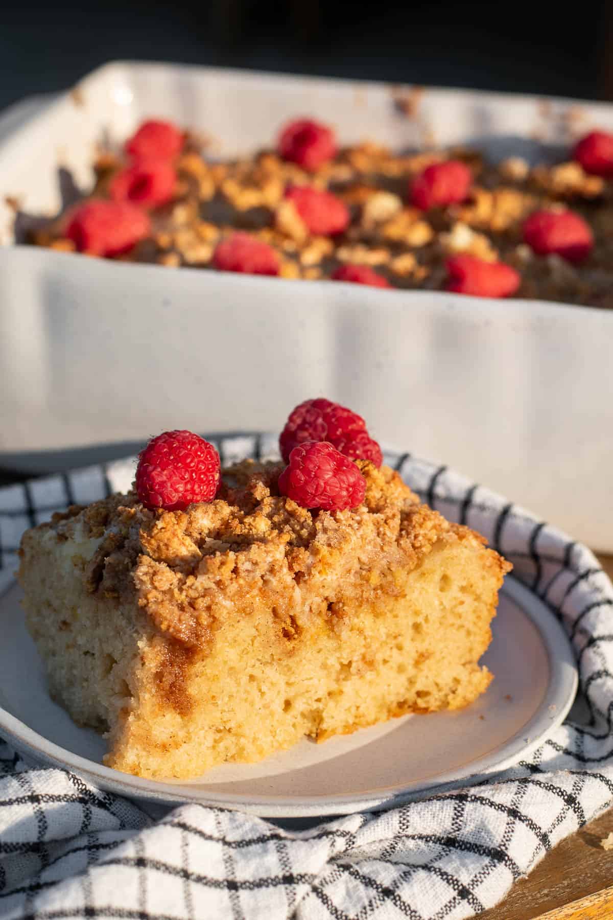 A large piece of focaccia topped with cheesecake filling, crushed graham crackers, and fresh raspberries sitting on a plate in the sunlight.