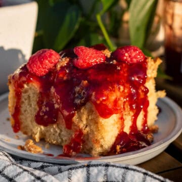 A large square of cheesecake focaccia topped with red jam and fresh raspberries next to a black and white linen.
