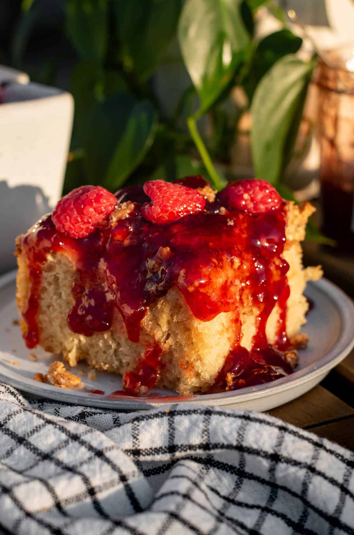A large square of cheesecake focaccia topped with red jam and fresh raspberries next to a black and white linen.