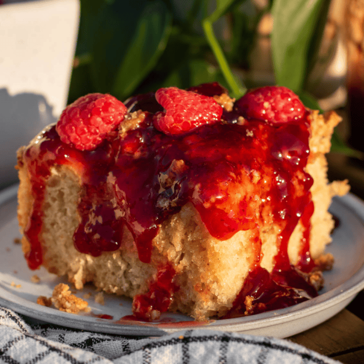 A large piece of focaccia topped with crushed graham crackers, cheesecake filling, strawberry jam, and fresh raspberries sitting on a plate in the sunlight.
