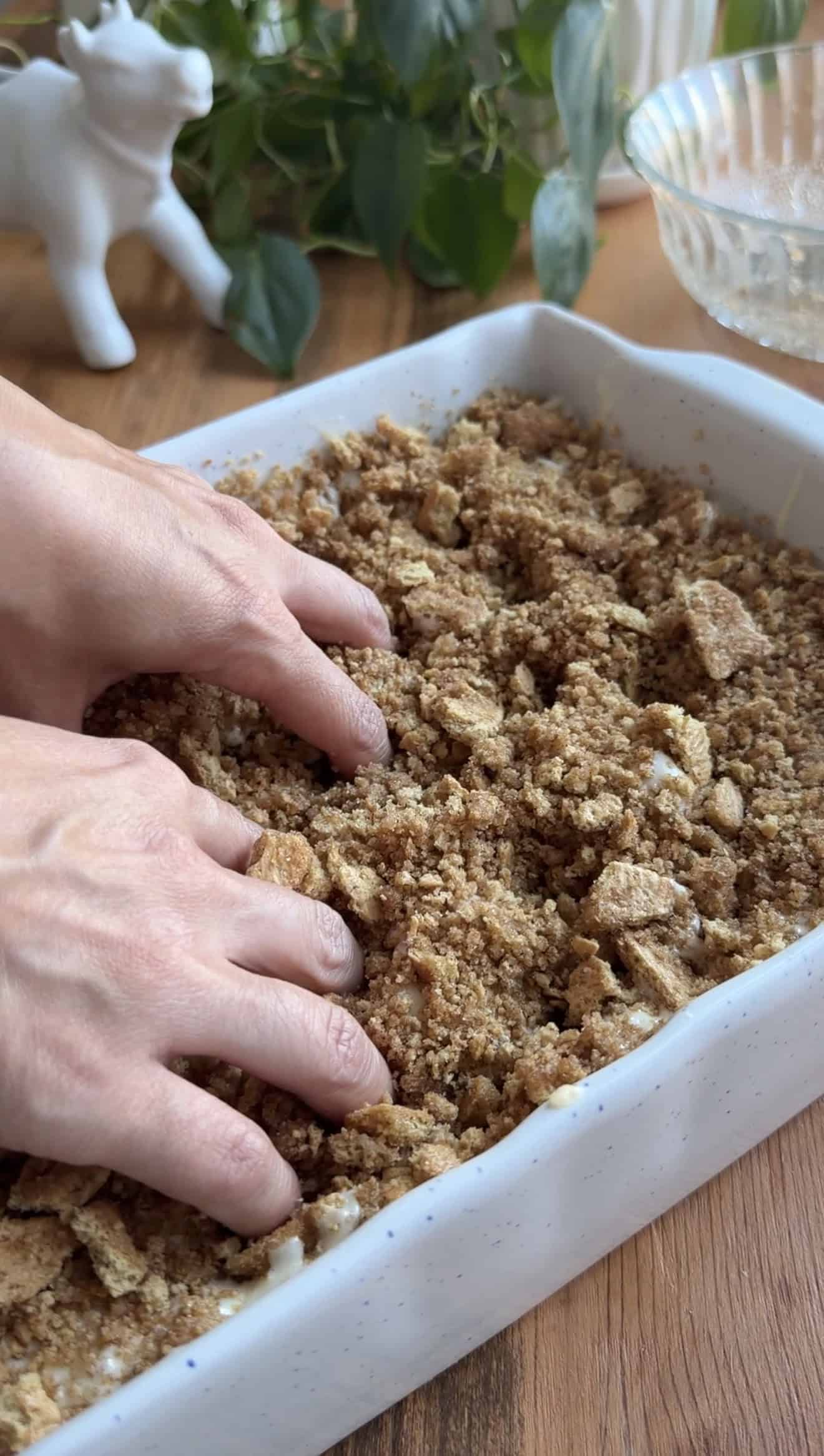 Two hands dimpling focaccia dough topped with crushed graham crackers in a white baking dish.