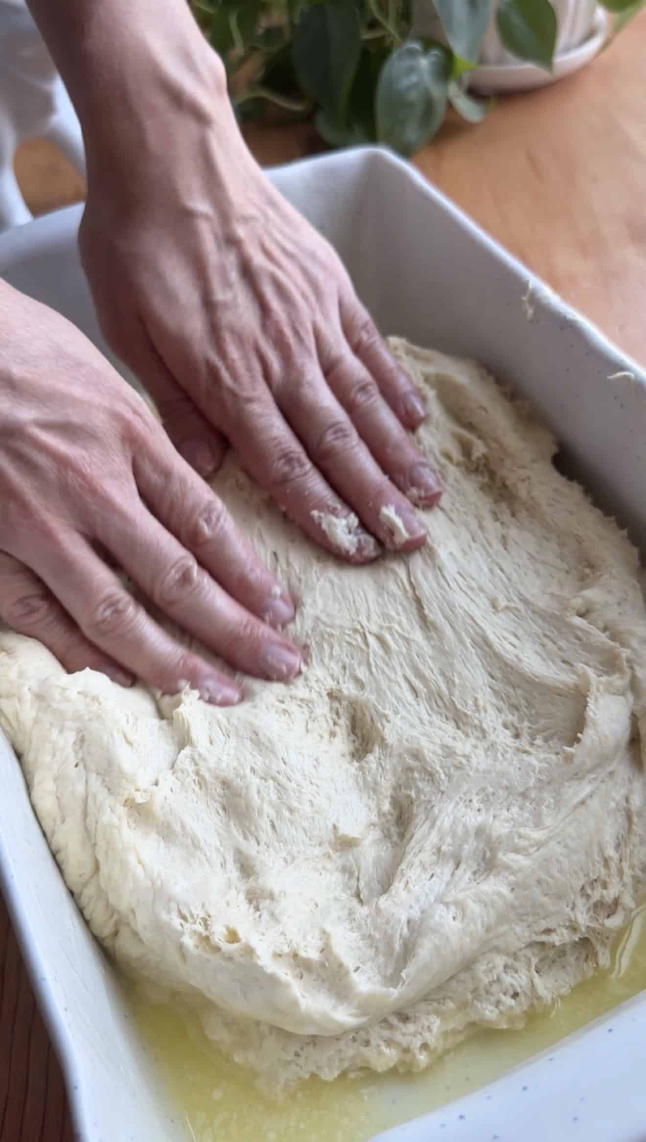 Two hands pressing bread dough into a baking dish greased with melted butter.