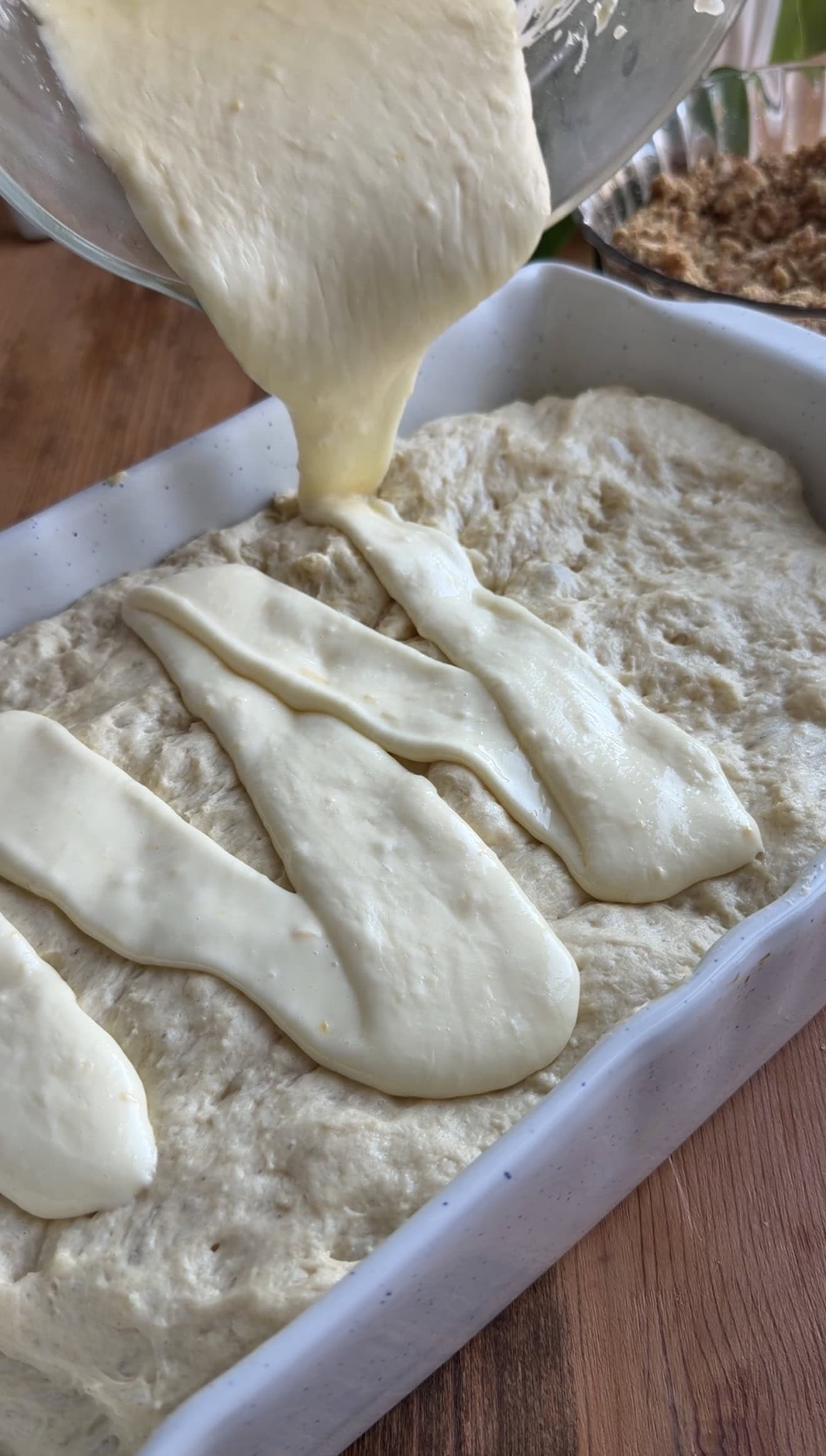A creamy cream-cheese-based mixture being poured from a glass mixing bowl onto bubbly focaccia dough in a white baking dish.
