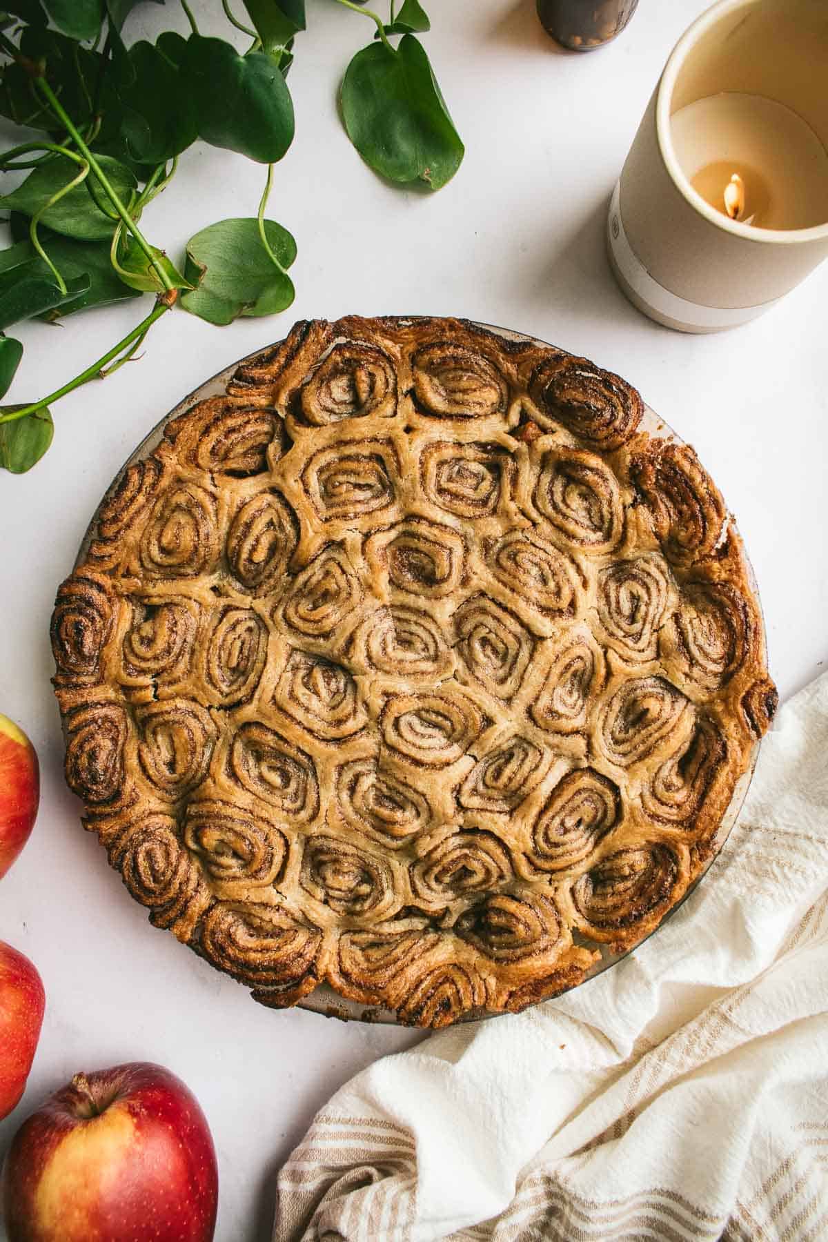 A golden, baked cinnamon roll apple pie next to whole fresh apples, a candle, and a kitchen towel.