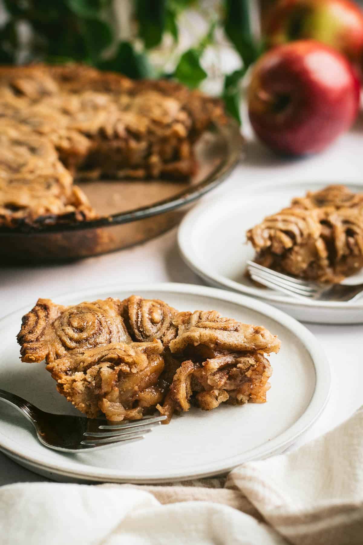 Two slices of cinnamon roll apple pie on grey plates next to the rest of the apple pie in a glass pie dish.