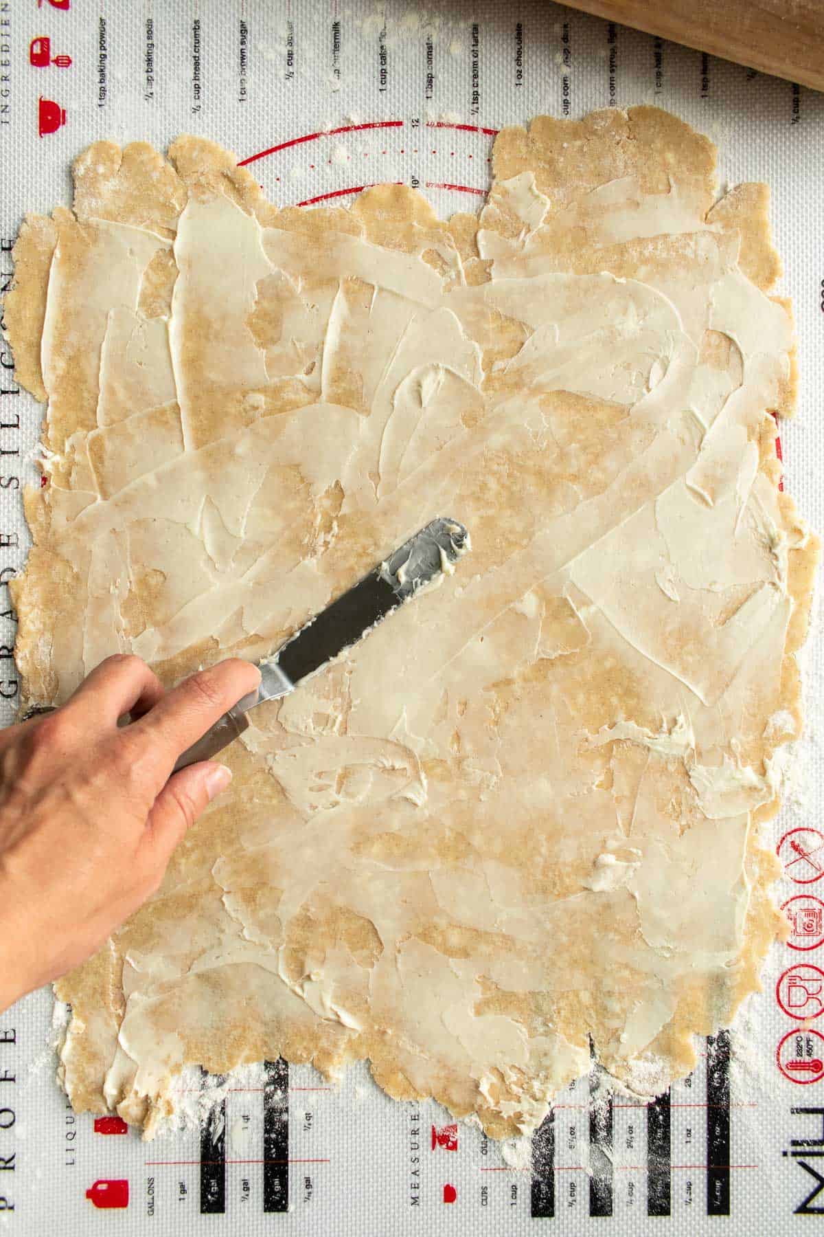 A hand spreading softened butter onto rolled-out pie crust.