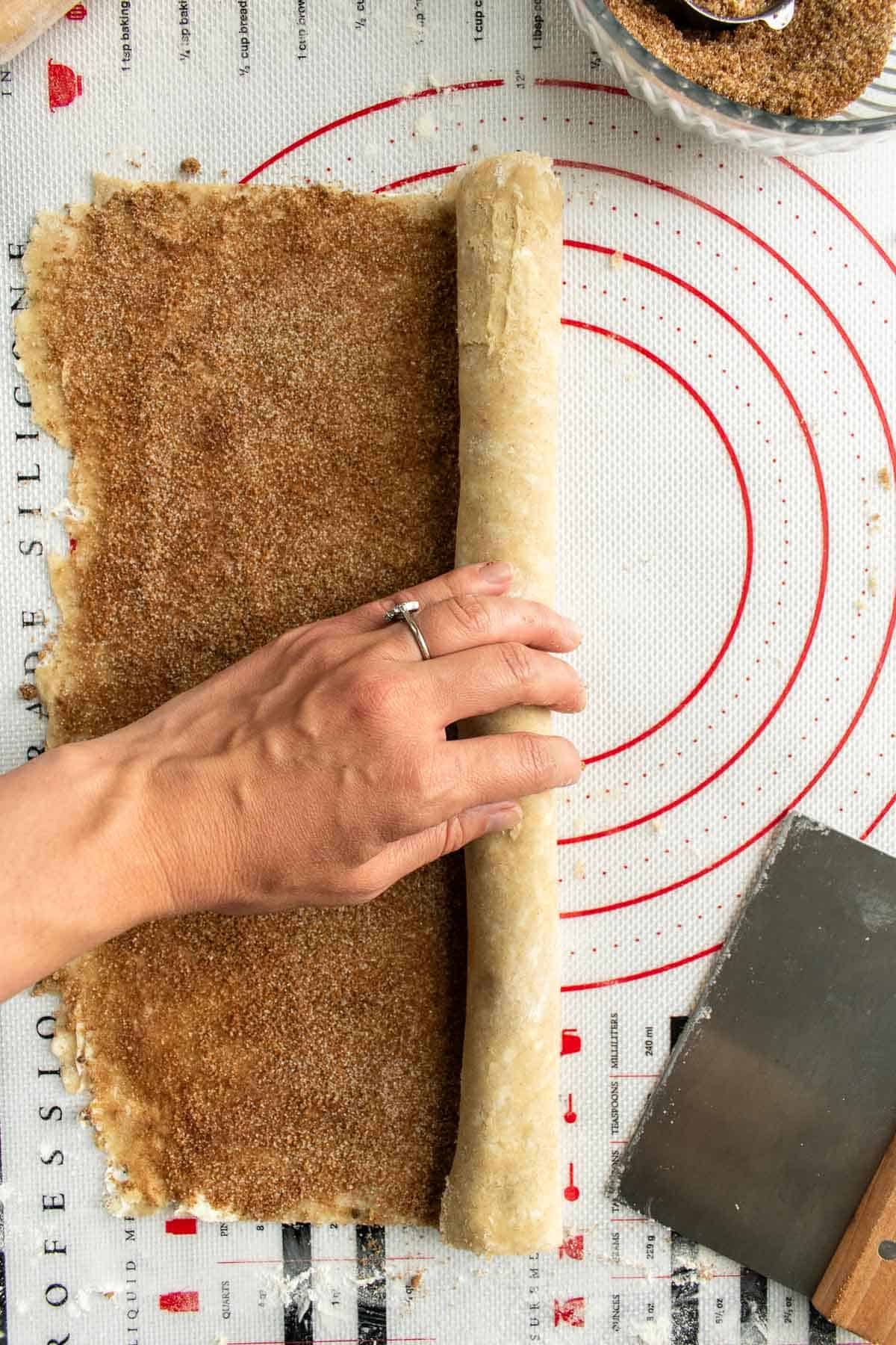 A hand rolling up a long log of pie crust topped with cinnamon sugar.