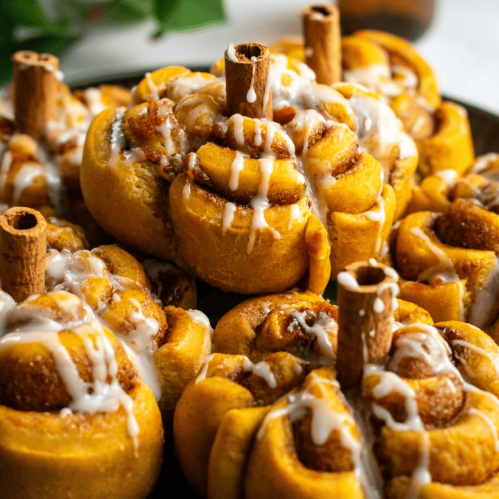 A stack of orange cinnamon rolls shaped like pumpkins topped with cinnamon sticks and vanilla glaze.