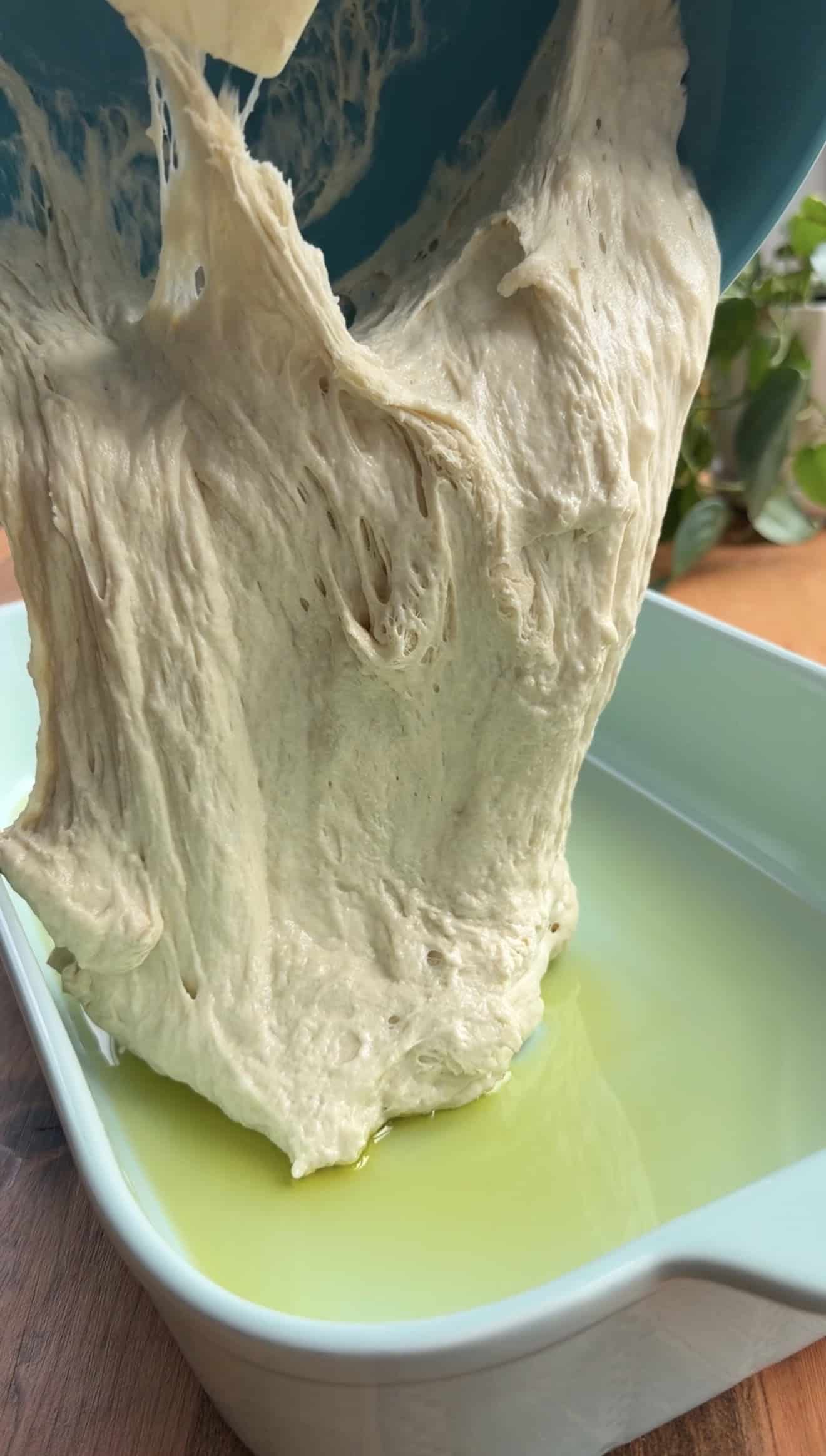 Bubbly, sticky bread dough being poured from a mixing bowl into an oiled baking dish.