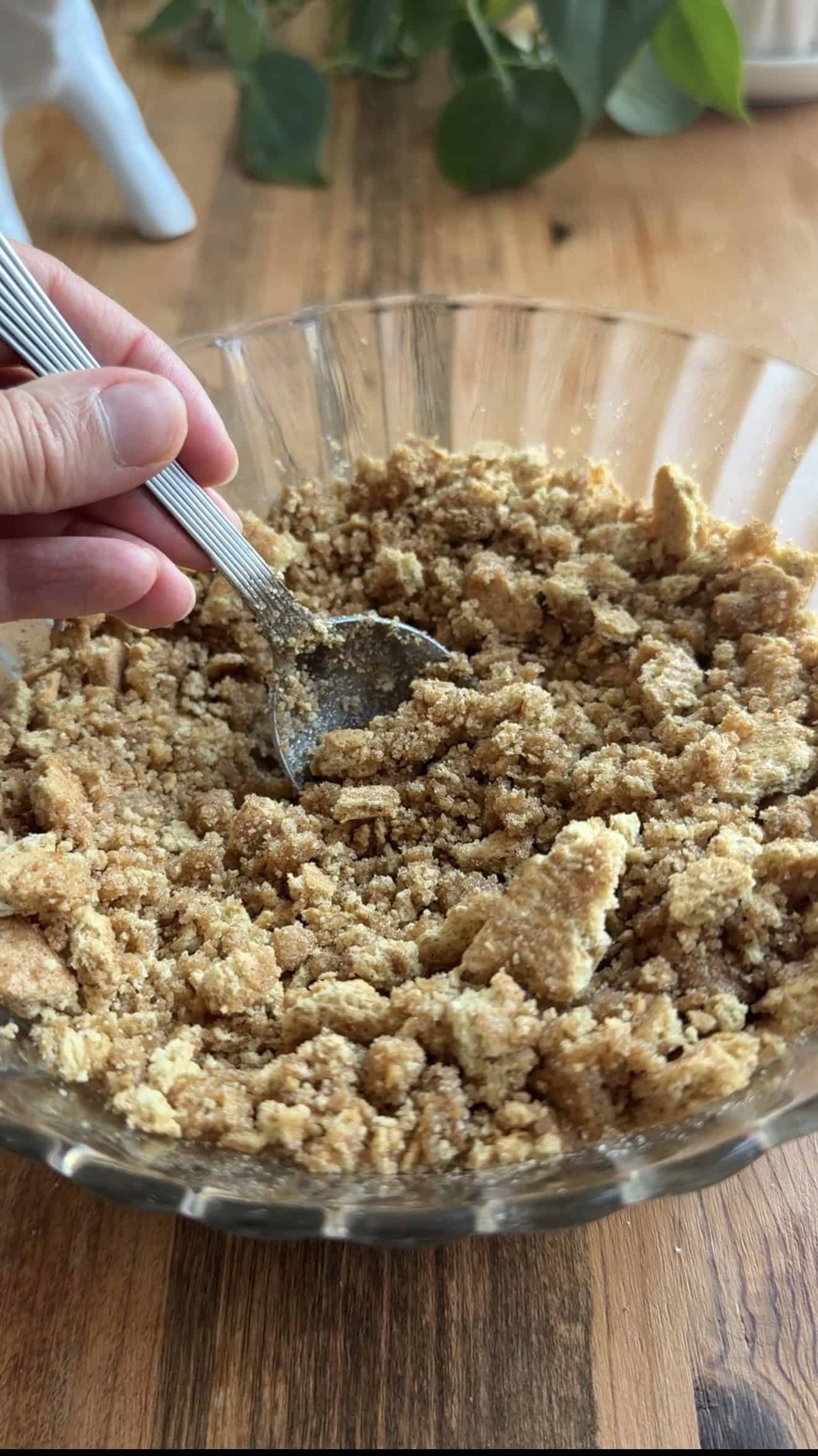A hand mixing crushed graham crackers with sugar and melted butter in a glass bowl.