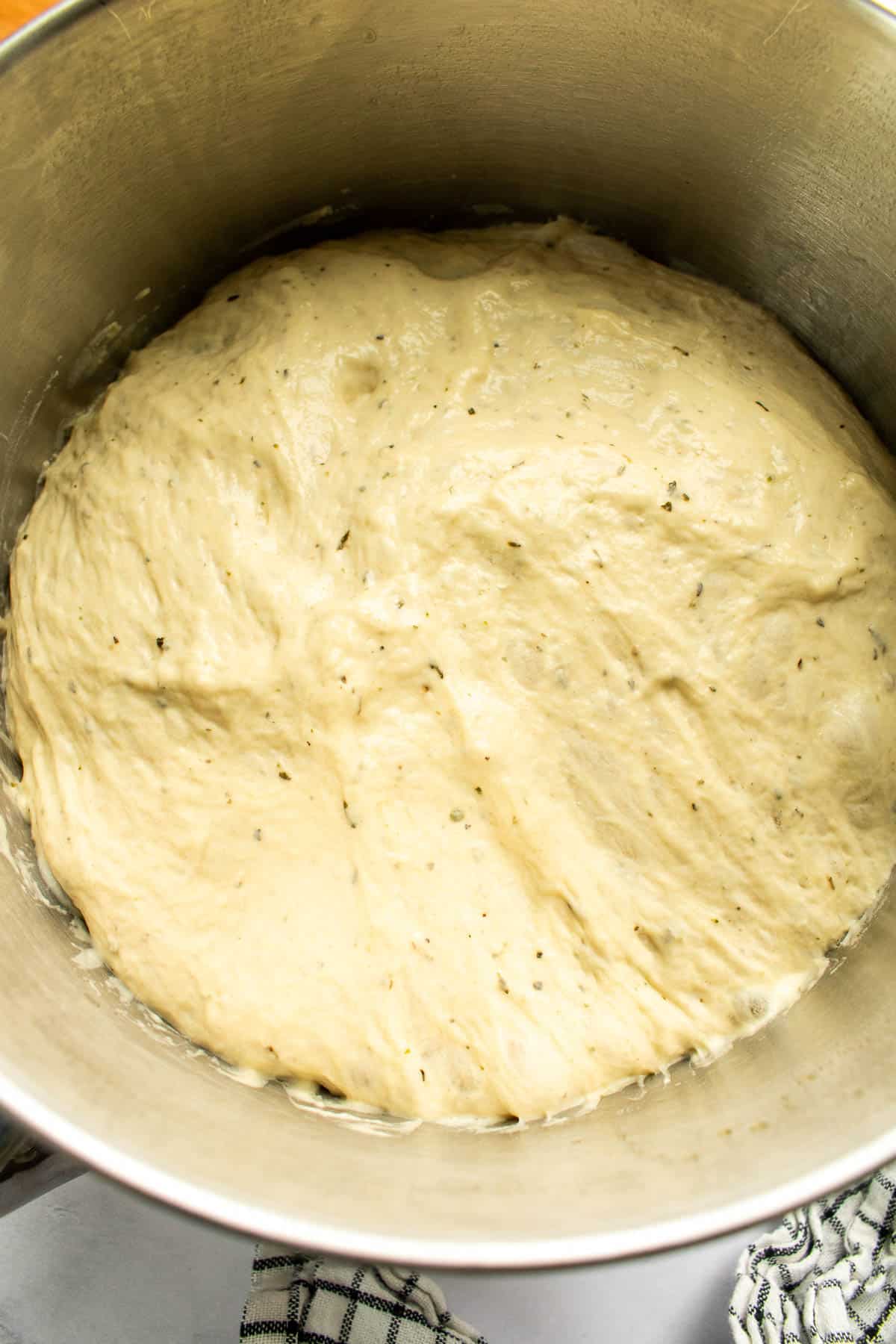 Risen bread dough with herbs mixed into it in a large metal bowl.