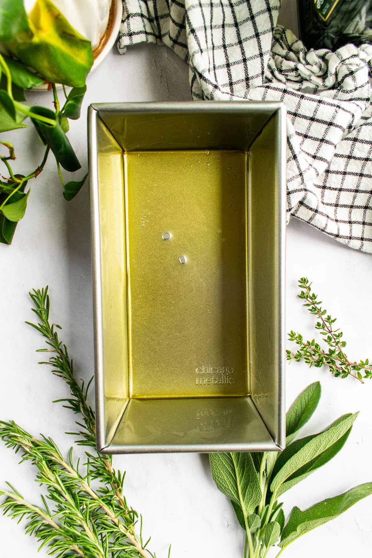 A metal loaf pan greased with olive oil next to fresh herb and a black and white linen.
