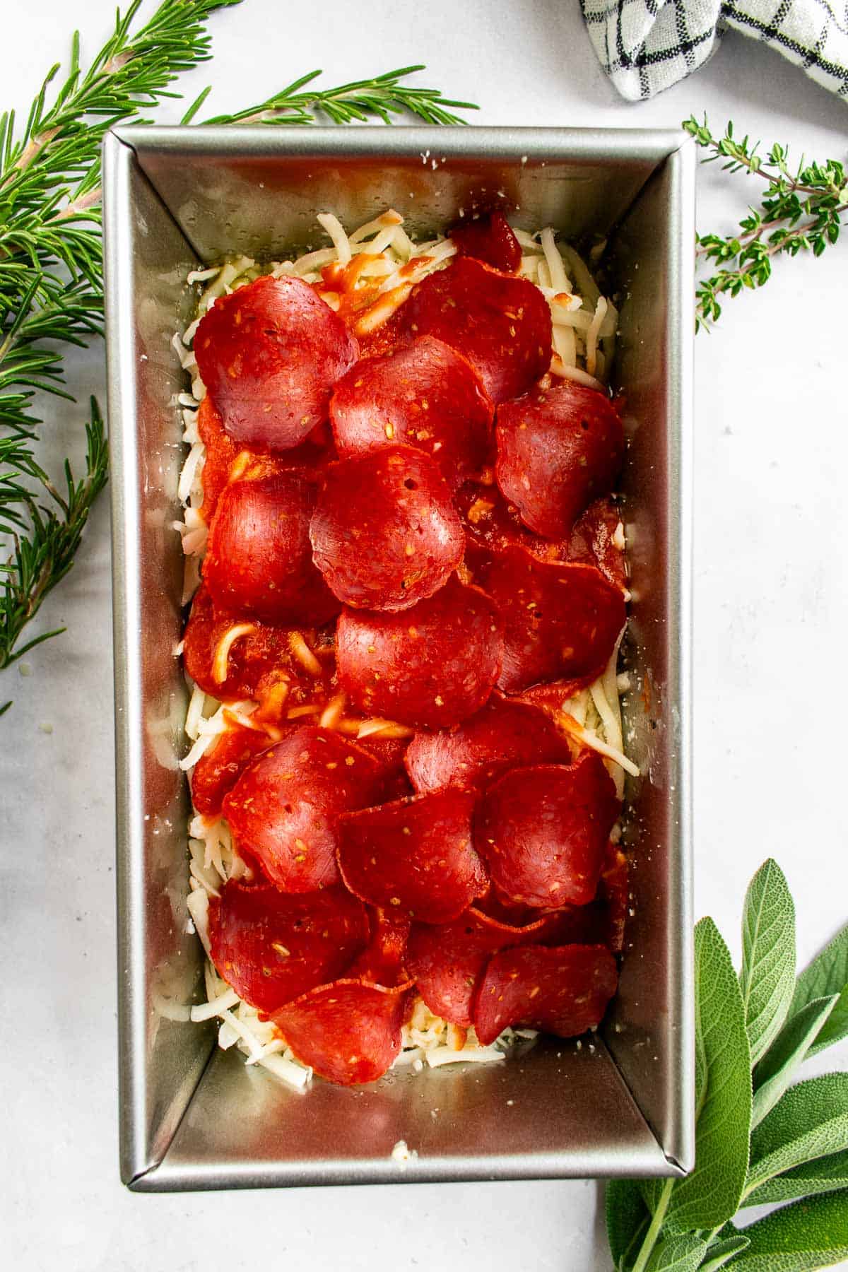 A metal loaf pan filled with focaccia dough topped with red sauce, shredded cheese, and pepperoni.