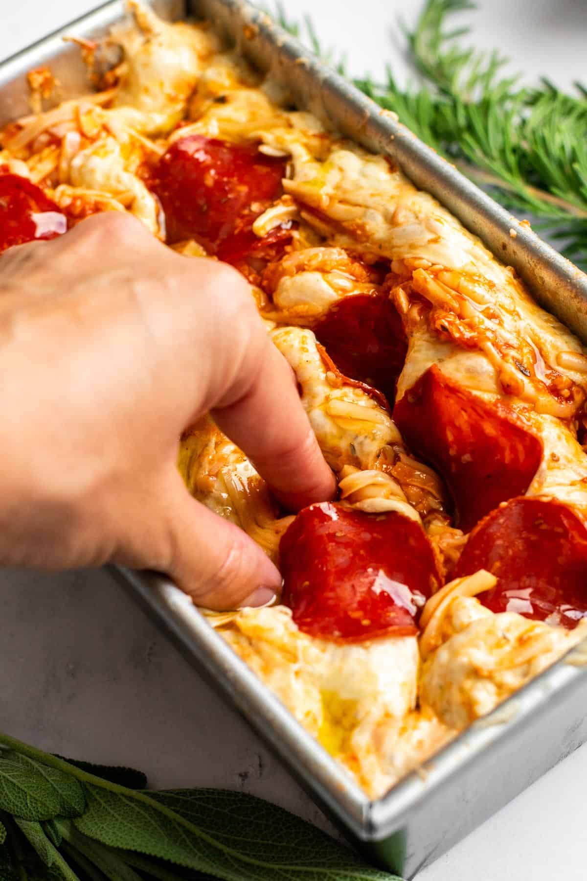 A hand dimpling focaccia dough in a loaf pan next to fresh herbs on a light surface.