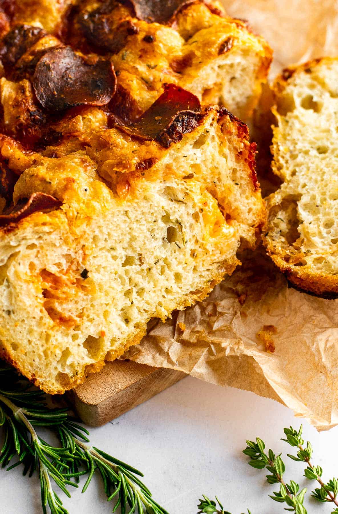 A slice of thick focaccia topped and filled with cheese and pepperoni next to fresh herbs on a light surface.