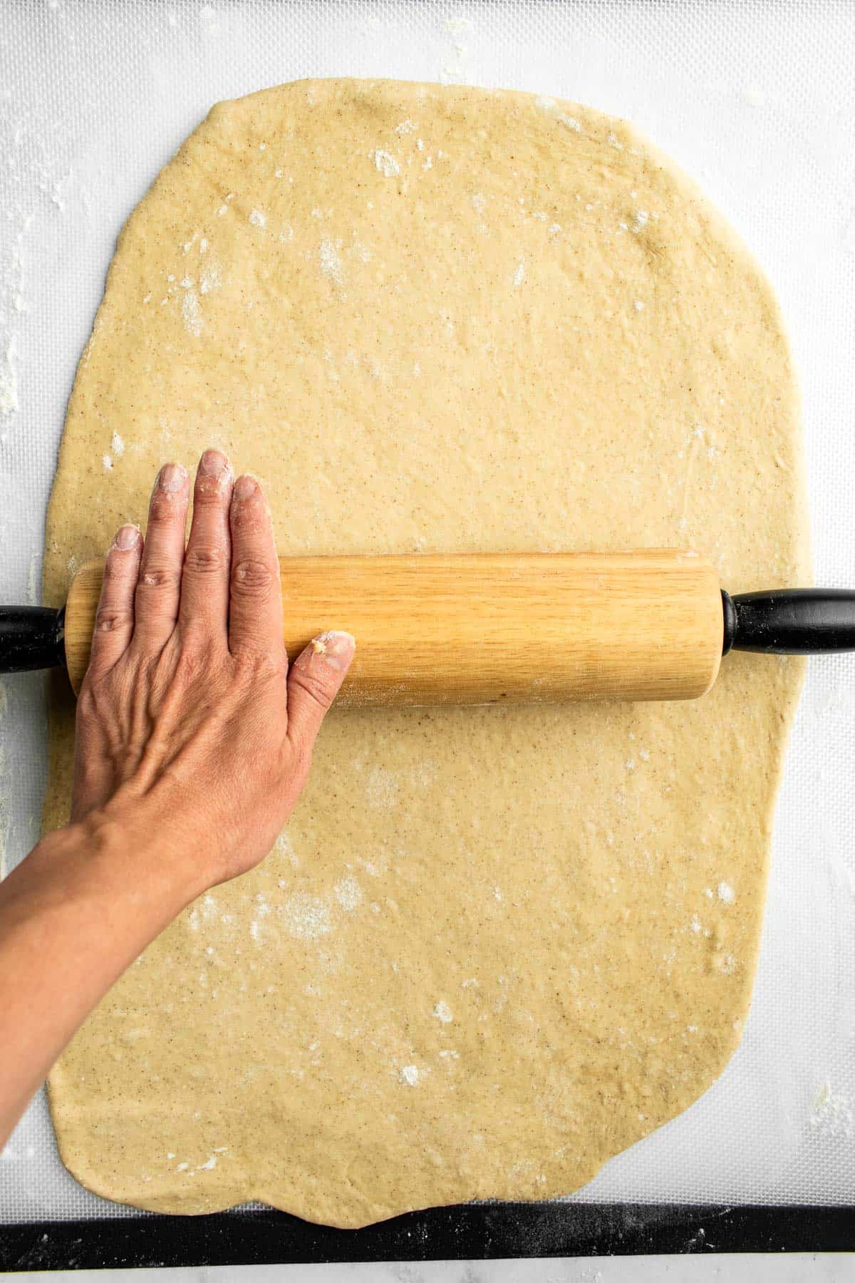 A hand rolling out cinnamon roll dough on a light surface with a wooden rolling pin.