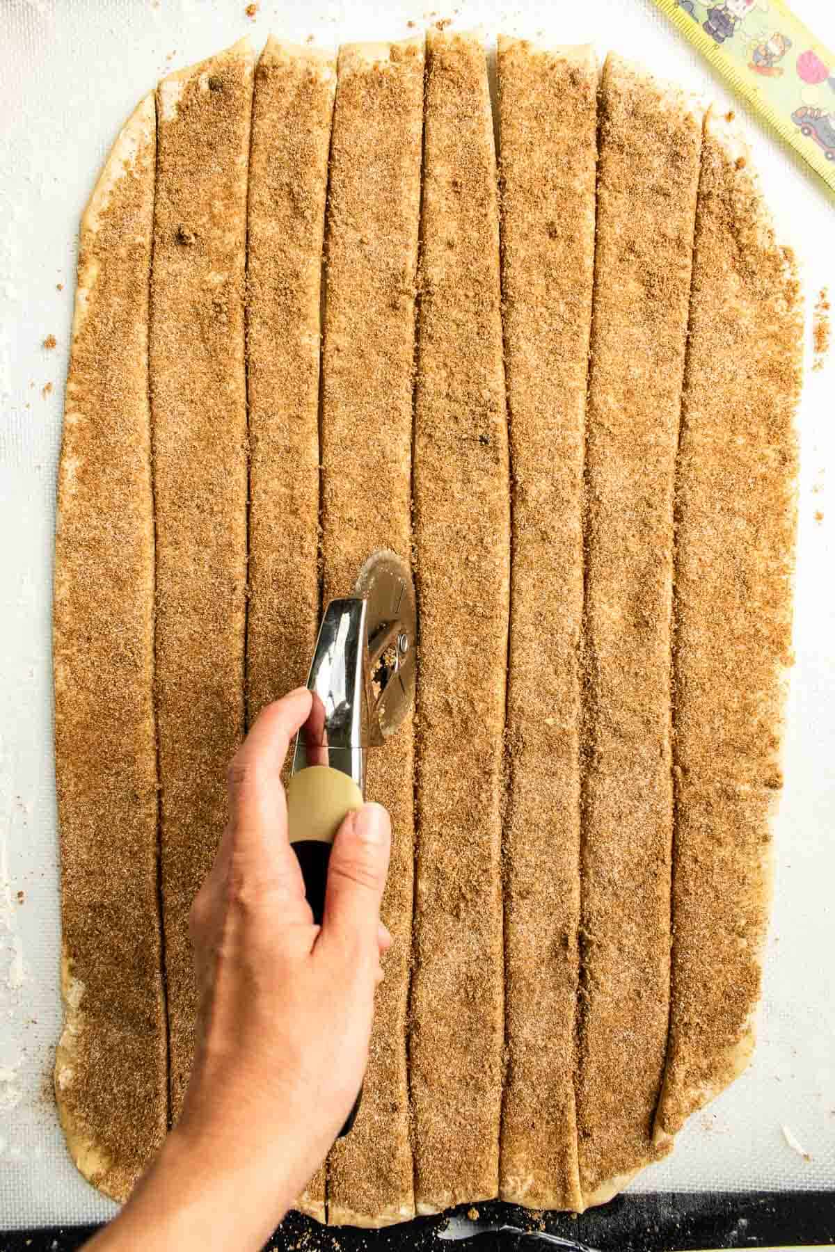 A hand cutting strips of cinnamon-sugar-coated bread dough with a pizza cutter.