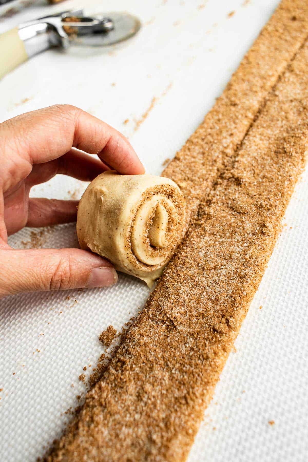 A hand rolling up a strip of cinnamon sugar-coated bread dough into a spiral.