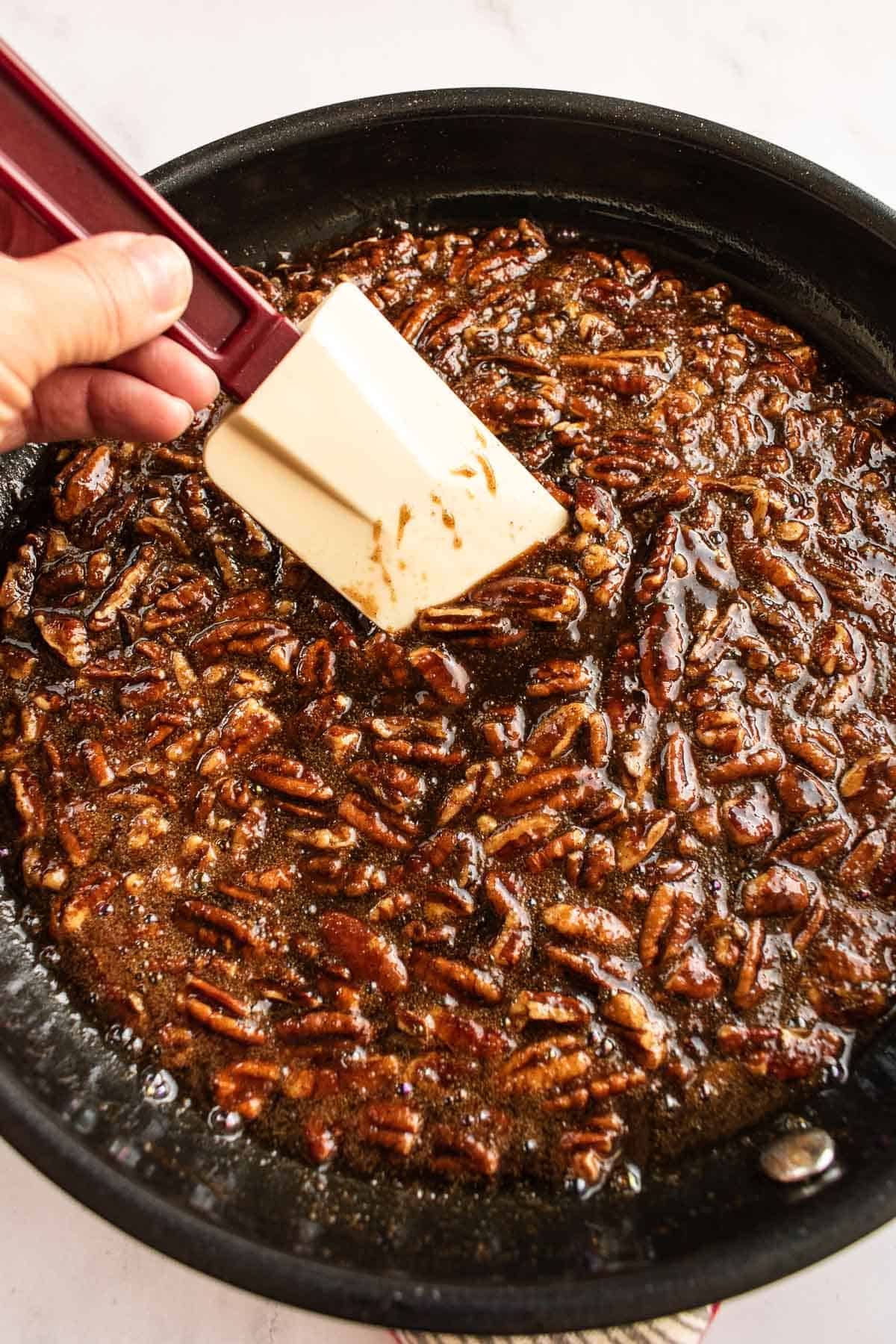 A hand stirring caramelized brown sugar, butter, and pecans in a saucepan with a silicone spatula.