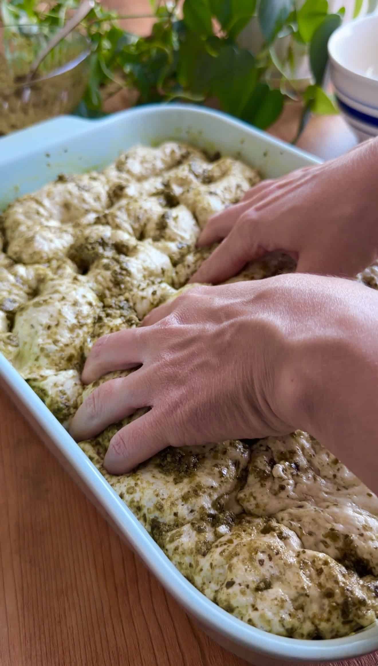 Two hands dimpling indentations into puffy basil focaccia dough in a blue baking dish.