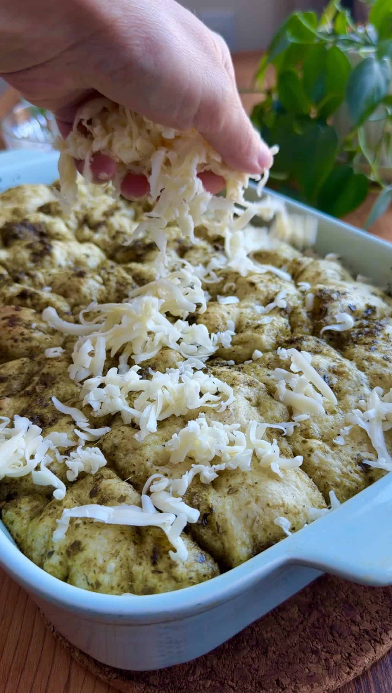 A hand sprinkling shredded white cheese onto golden focaccia in a baking dish that's coated in pesto.