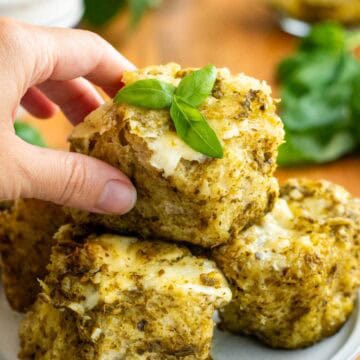 A hand holding a small piece of pesto focaccia topped with melted cheese and fresh basil leaves.