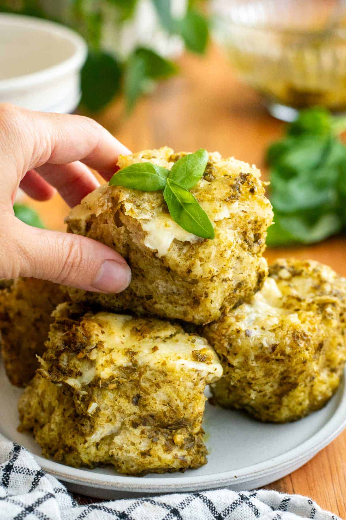 A hand holding a small piece of pesto focaccia topped with melted cheese and fresh basil leaves. 