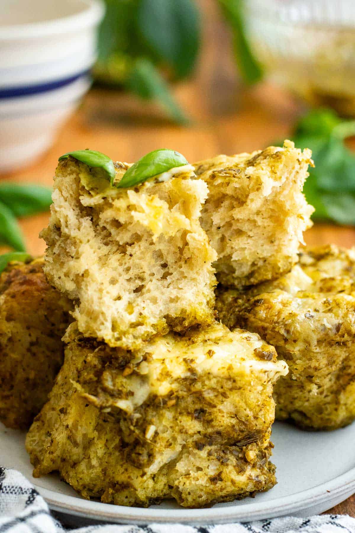 A piece of pesto focaccia torn in half to show the open crumb, stacked on top of more pieces of pesto focaccia on a grey plate.