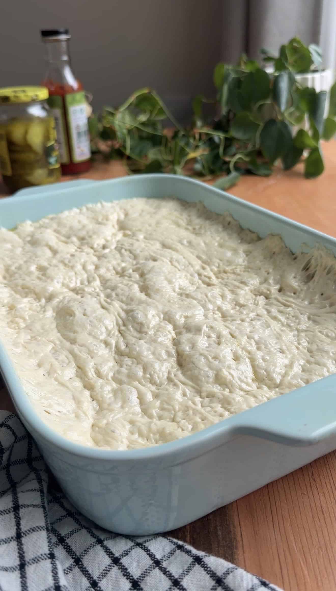 Bubbly, risen focaccia in a large blue baking dish on a table near a plant.