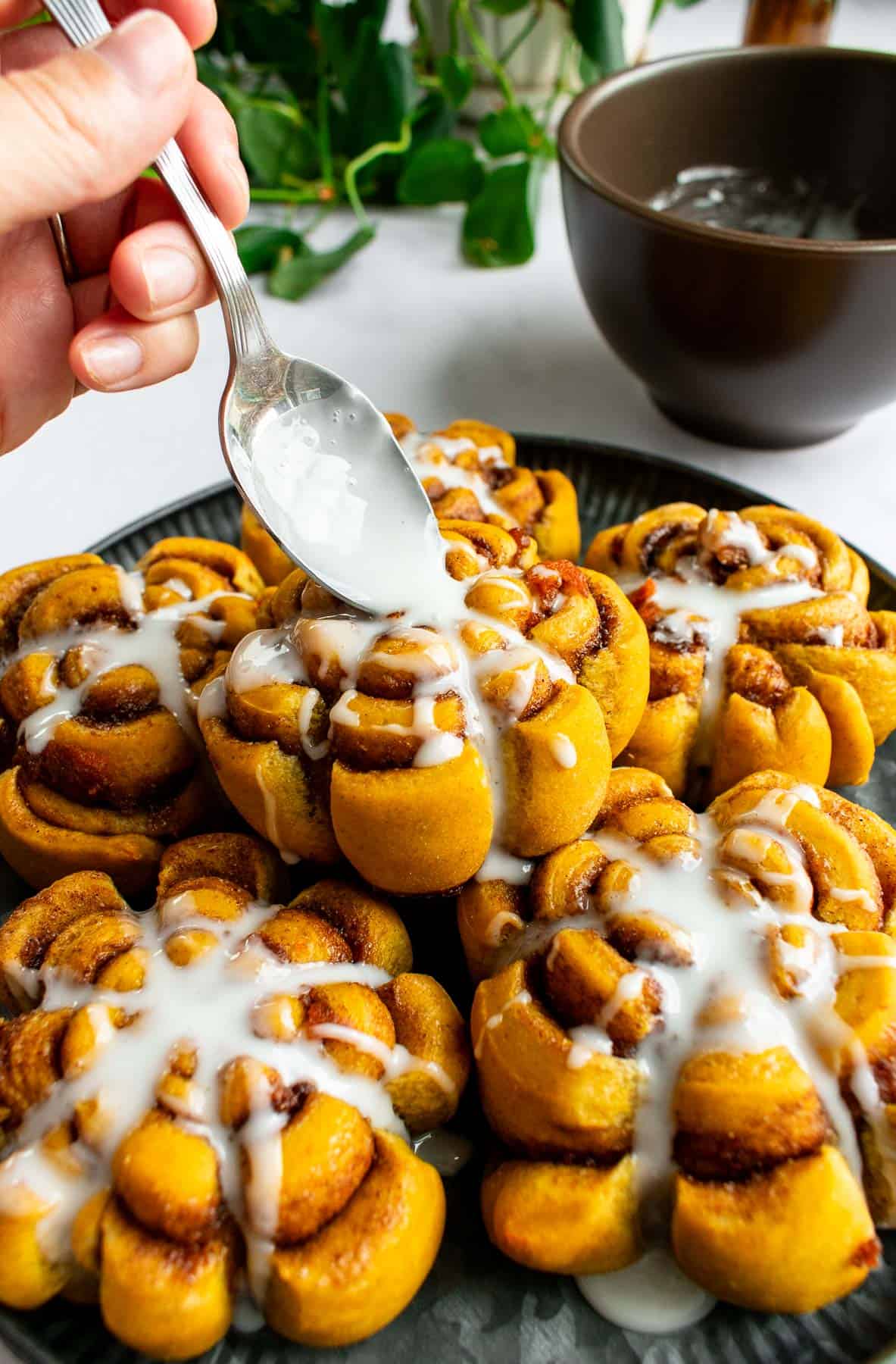 A hand drizzling white glaze from a spoon onto pumpkin cinnamon rolls.