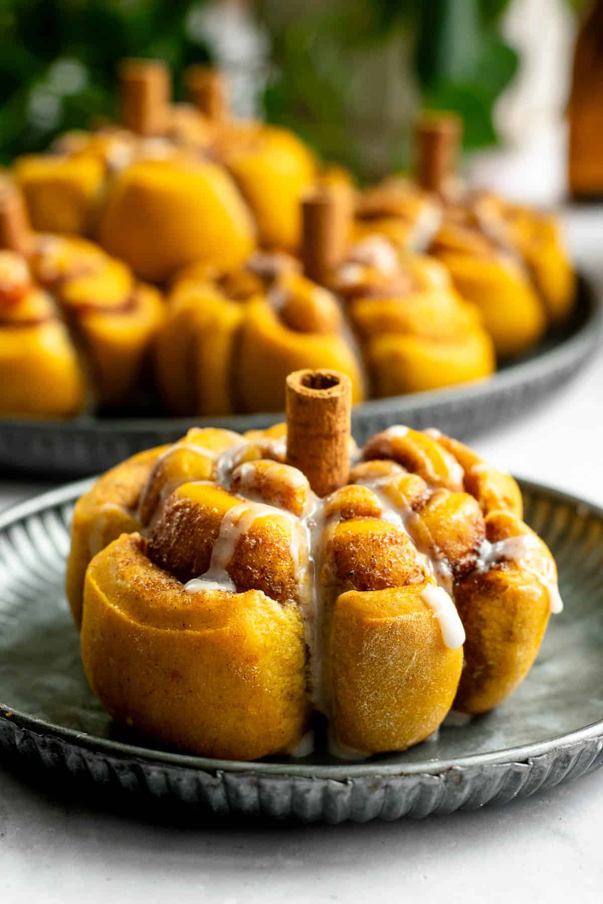 A large pumpkin cinnamon roll with a cinnamon stick stem, shaped like a pumpkin, sitting on a grey plate.