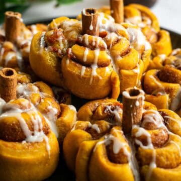 A metal plate topped with half a dozen glazed pumpkin-shaped cinnamon rolls with cinnamon stick stems.