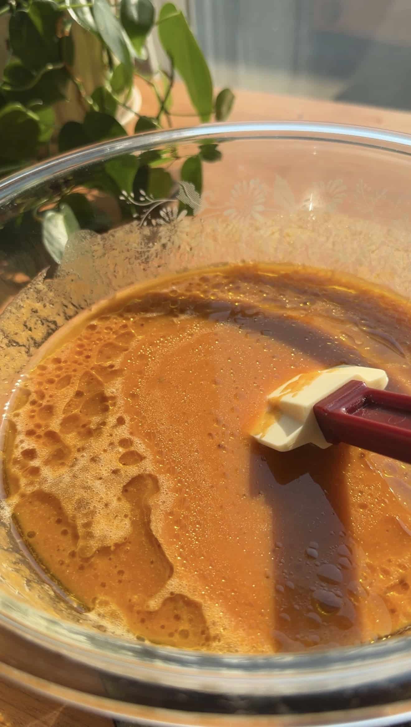 A silicone spatula sitting in a glass mixing bowl filled with orange liquid on a table.