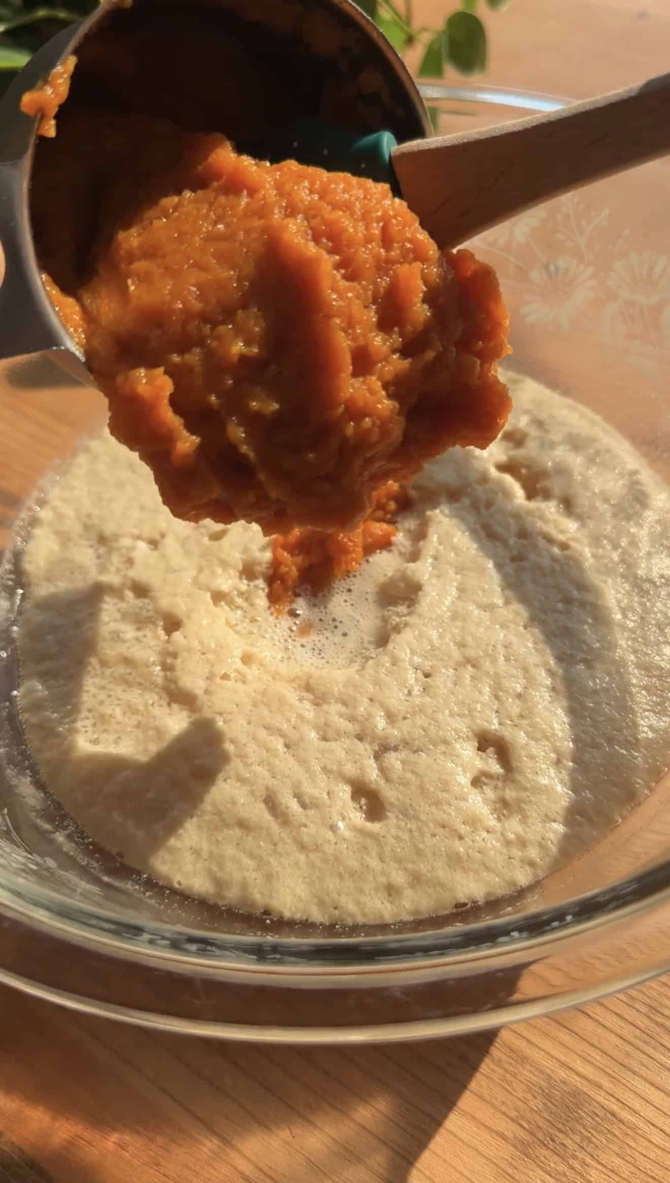 Pumpkin puree being poured from a measuring cup into a mixing bowl with frothy yeast and water.