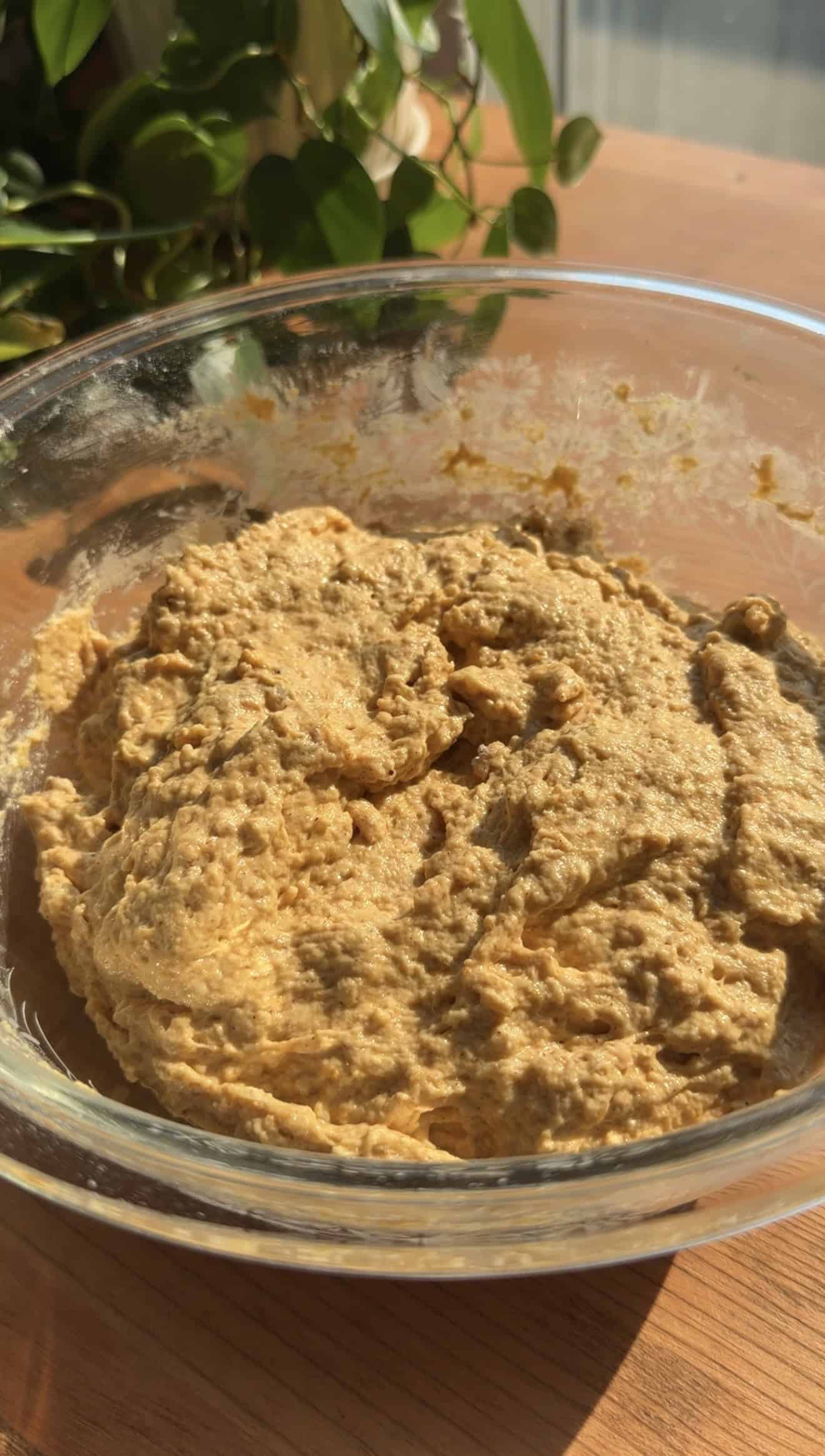 Light orange, sticky bread dough in a glass mixing bowl on a light brown table.